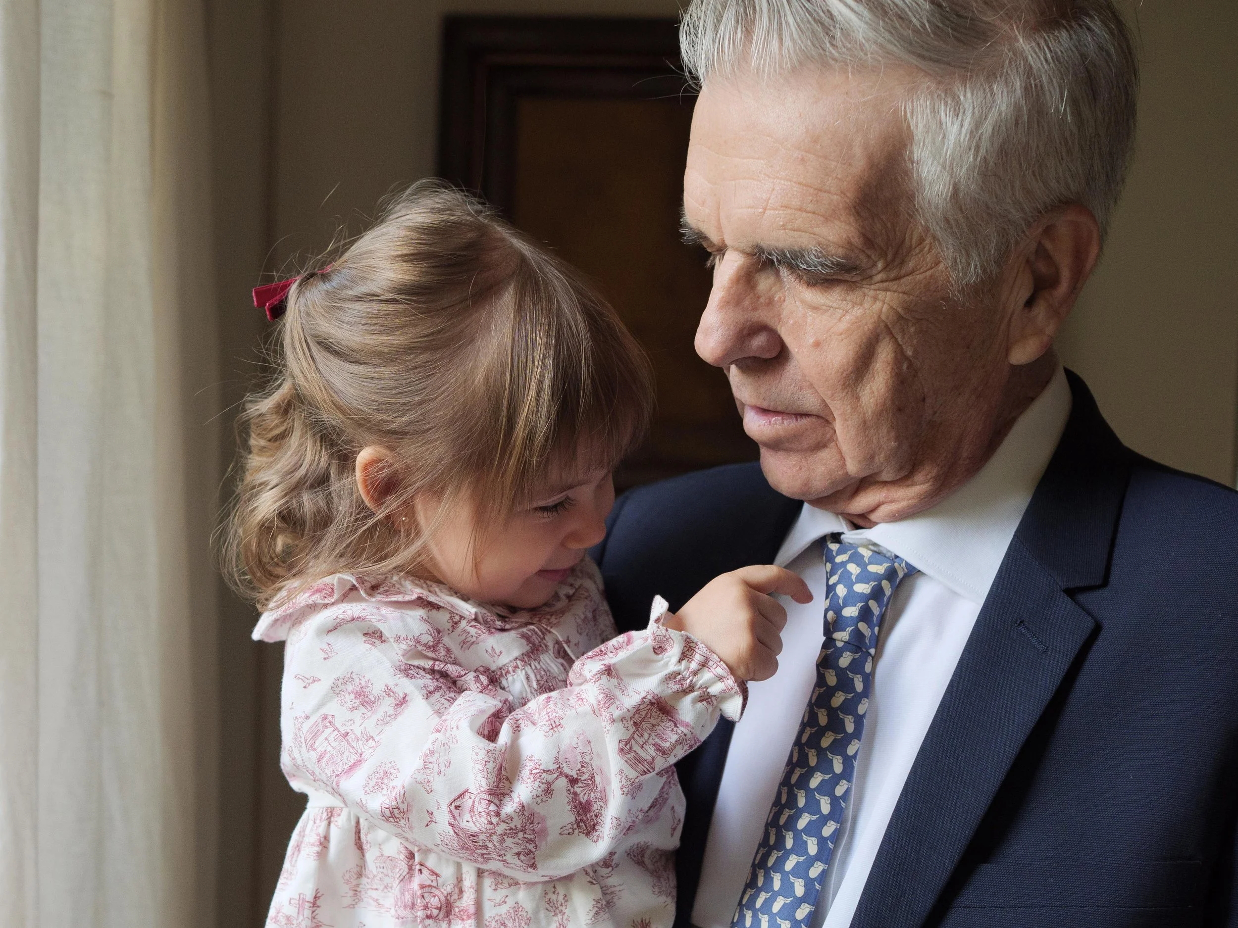 An elderly man in a formal suit is gently holding a young girl, who is touching his face affectionately, in a warmly lit room.