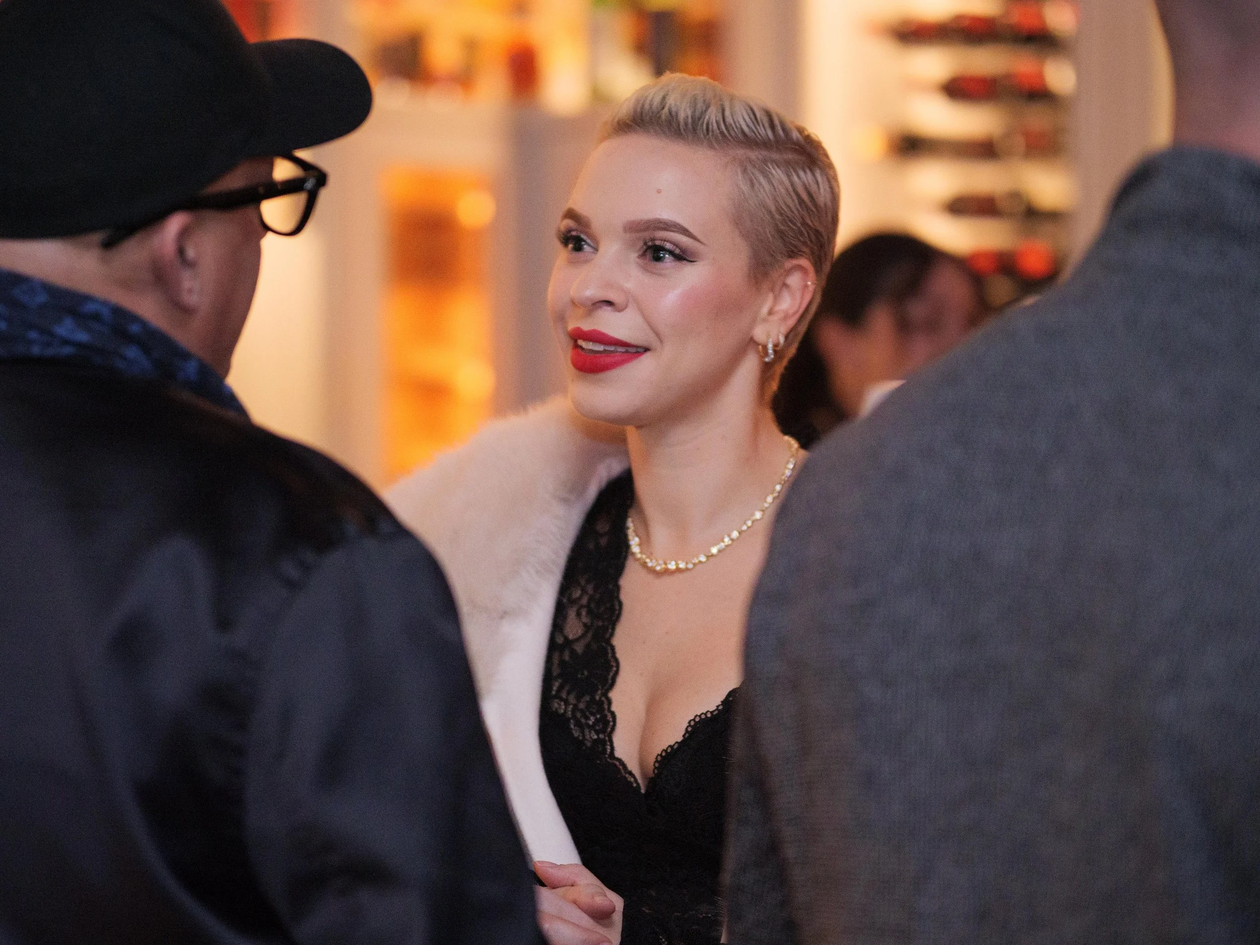 Woman with blonde hair, red lipstick, wearing a black lace dress, pearl necklace, and earrings, talking to men in a social setting.