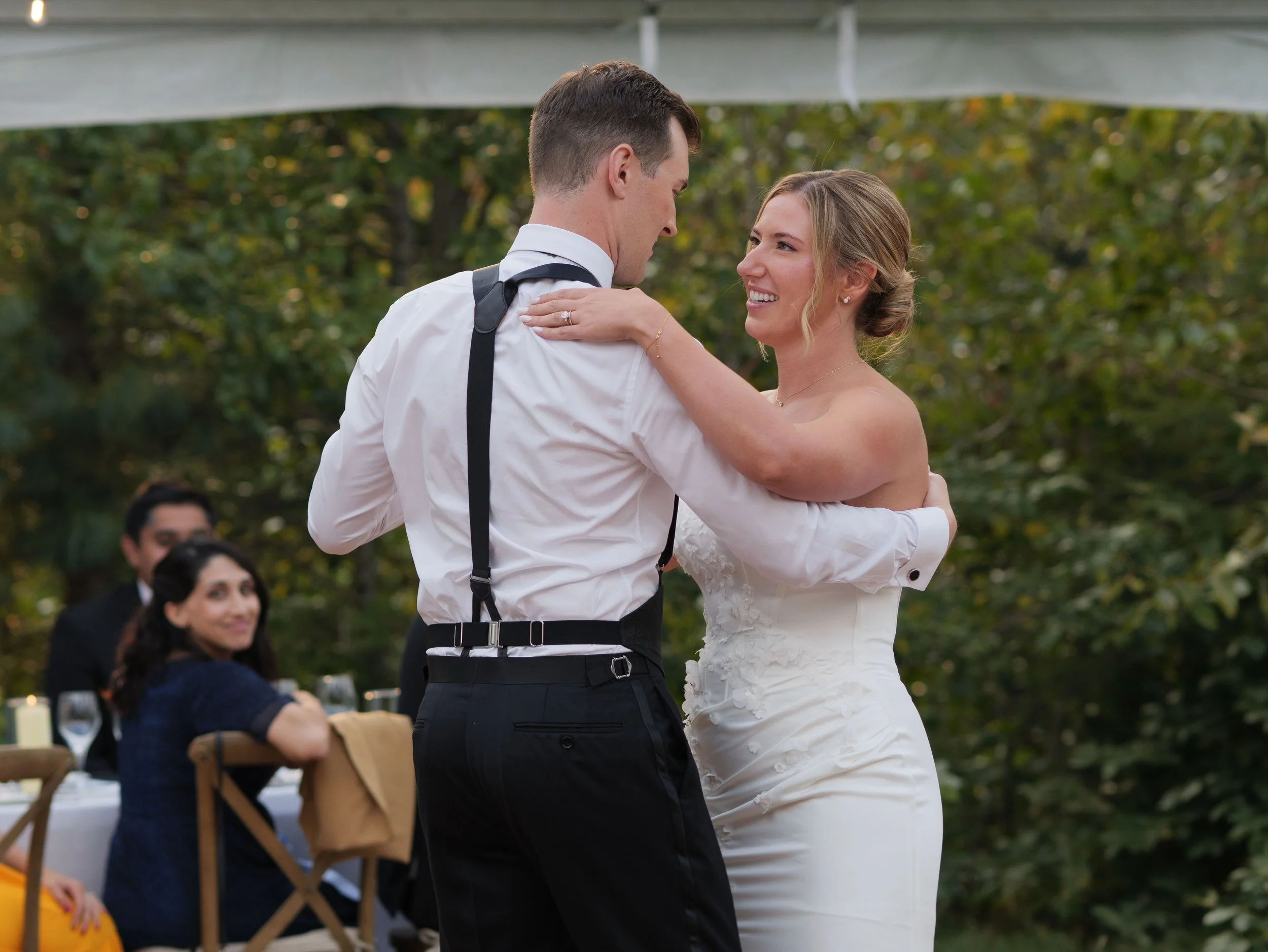 A couple dancing at their wedding reception outdoors, with guests watching and trees in the background.