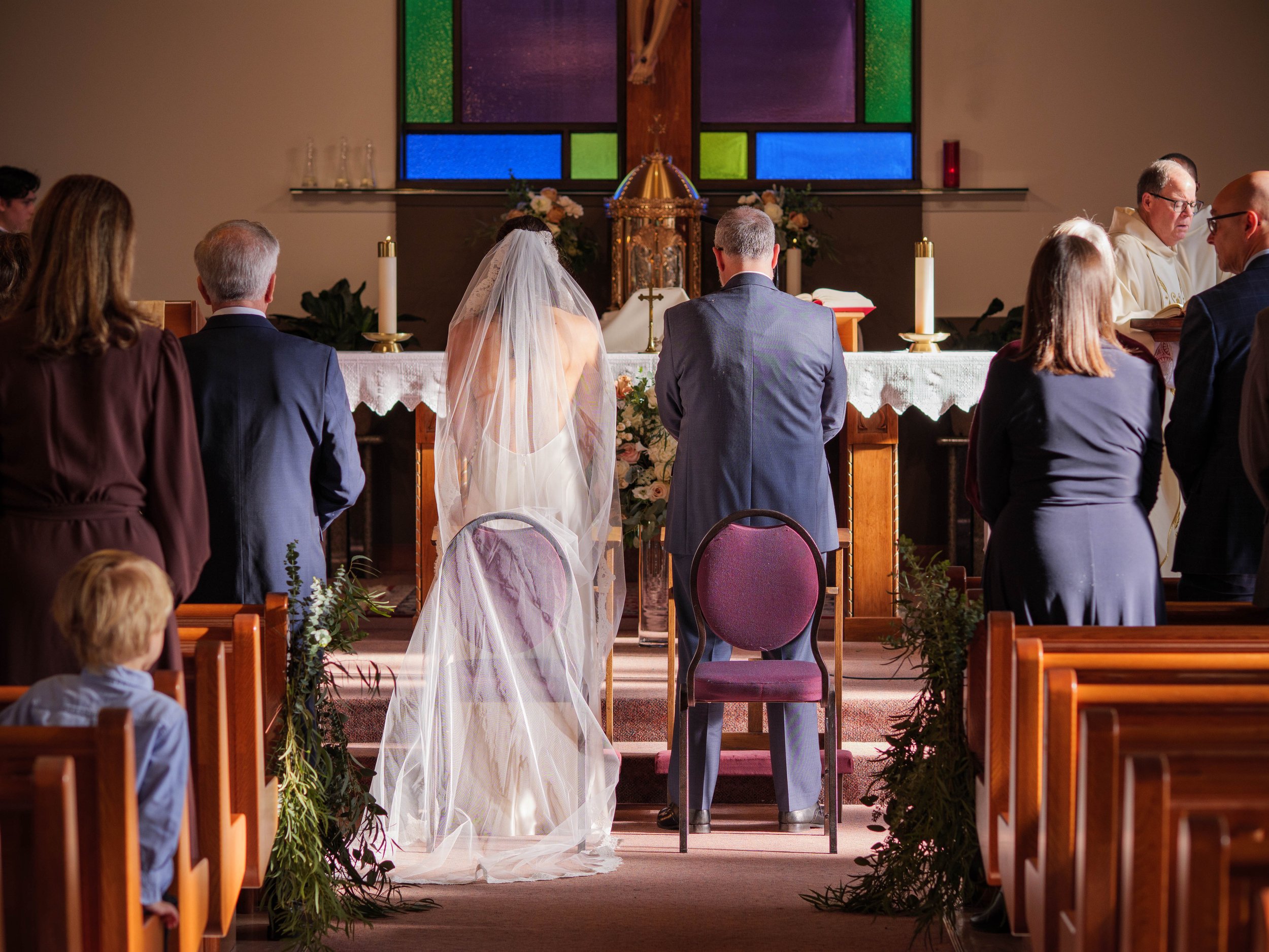 A wedding ceremony takes place inside a church with people standing at the altar, including a bride with a veil and a groom, and a priest conducting the service, in front of a stained glass window.