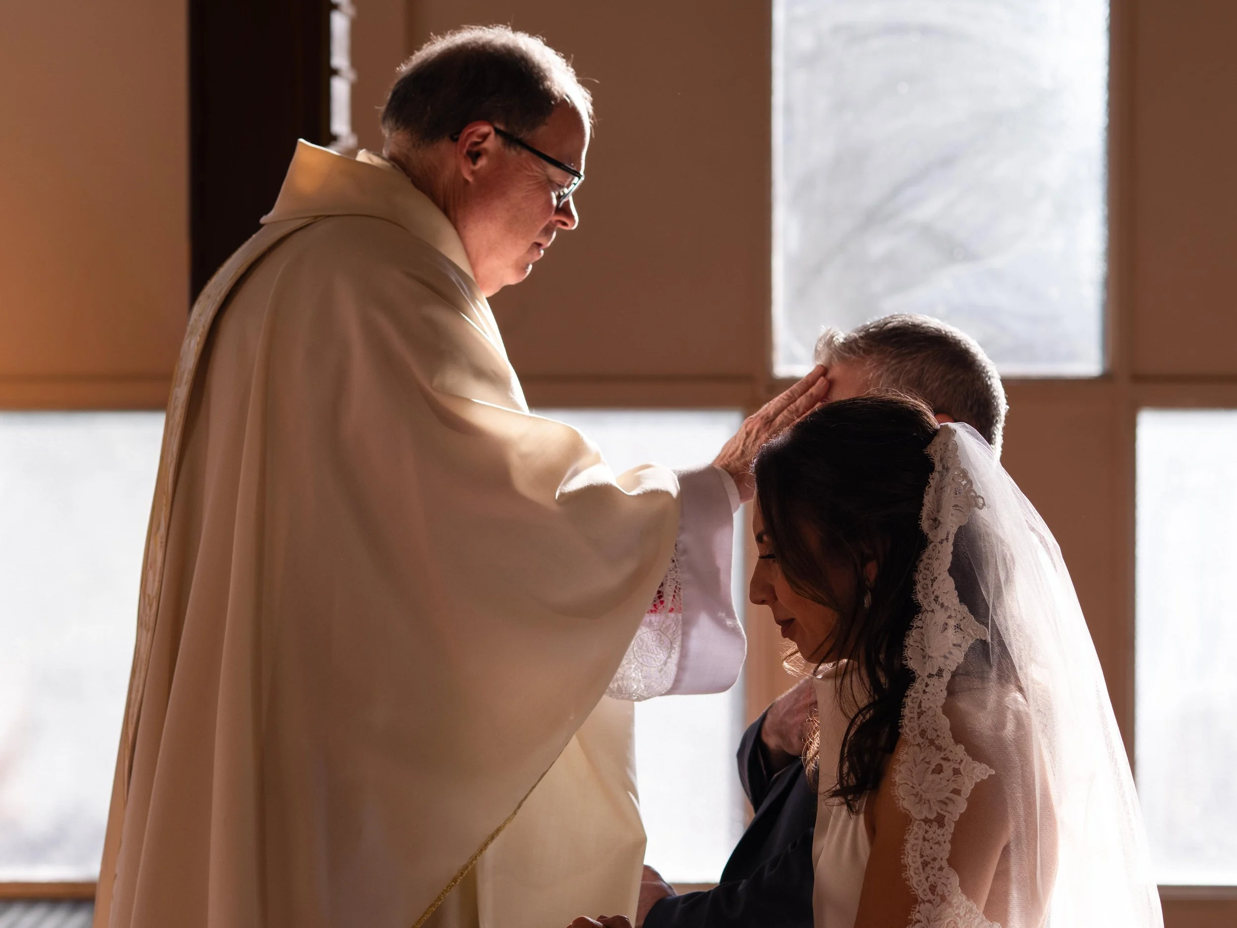 A priest performing a wedding ceremony, placing his hand on the forehead of a bride, with the groom kneeling beside her, inside a church with large windows letting in natural light.