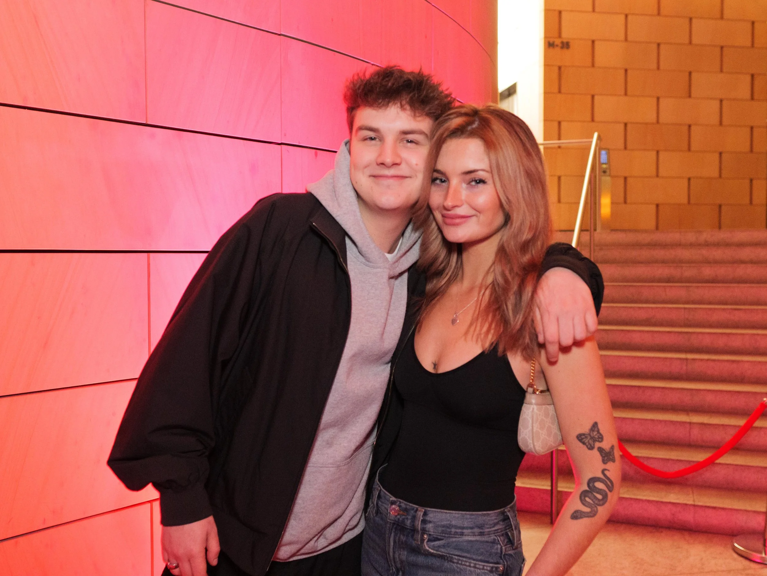 A young man and woman are standing close together on a staircase, smiling at the camera. The man is wearing a black jacket over a gray hoodie, and the woman is in a black top with tattoos on her arm. She has light brown hair and is holding a small pu