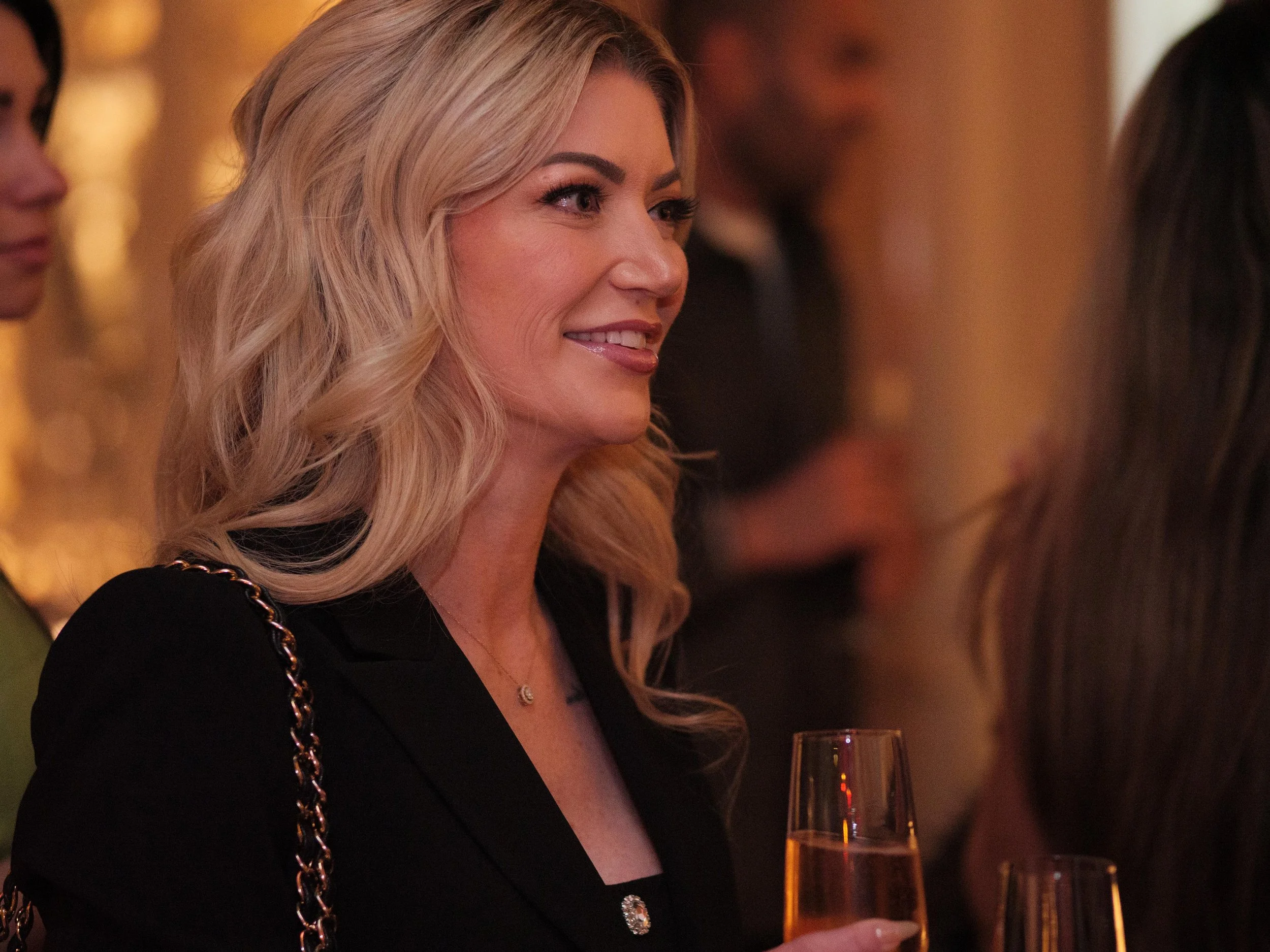 A blonde woman with wavy hair smiling and holding a glass of champagne at a social event.