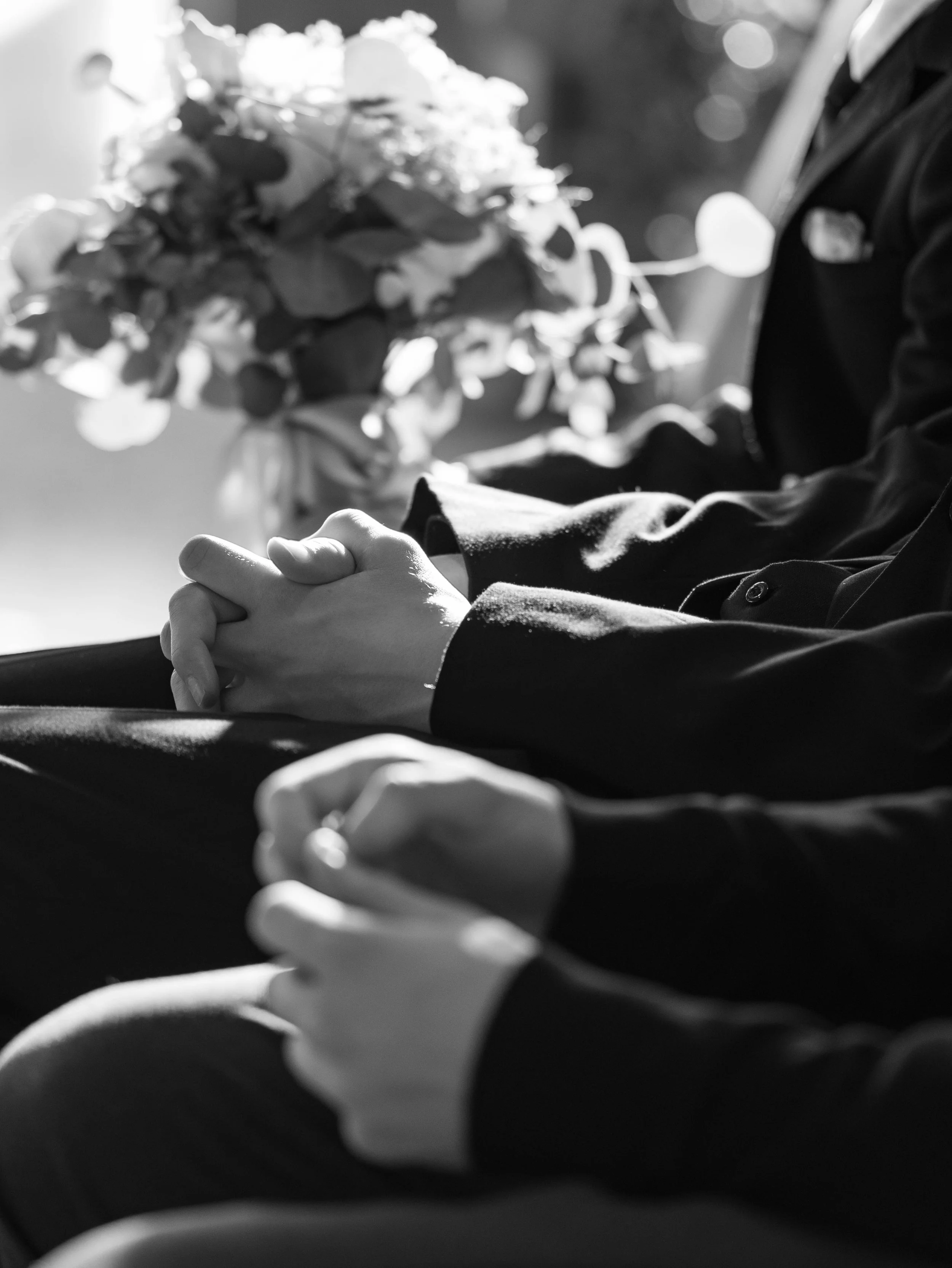 Close-up of two people holding hands, dressed in formal attire, with a blurred bouquet of flowers in the background.
