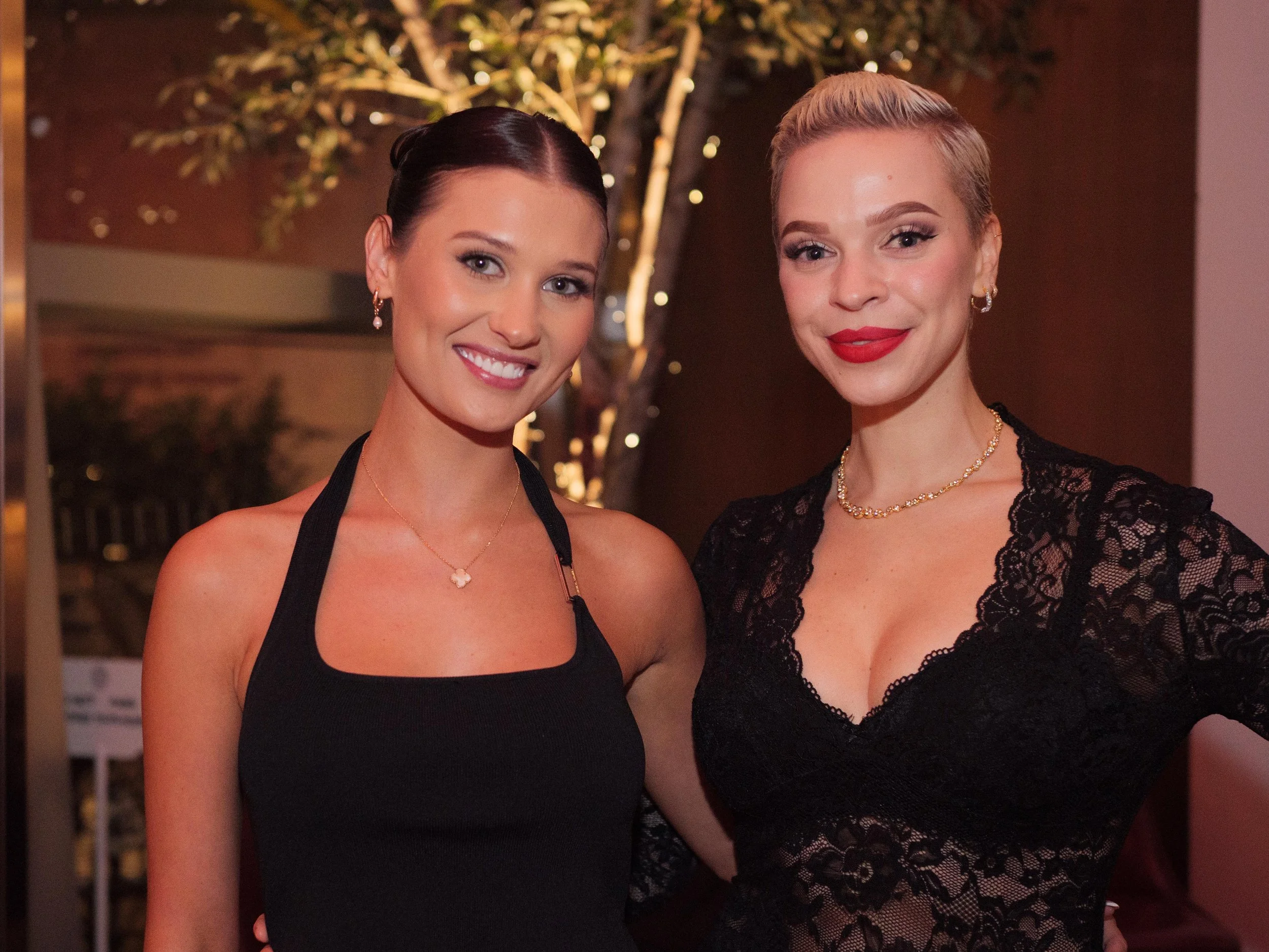 Two women dressed in black outfits smiling at an indoor event with trees and lights in the background.