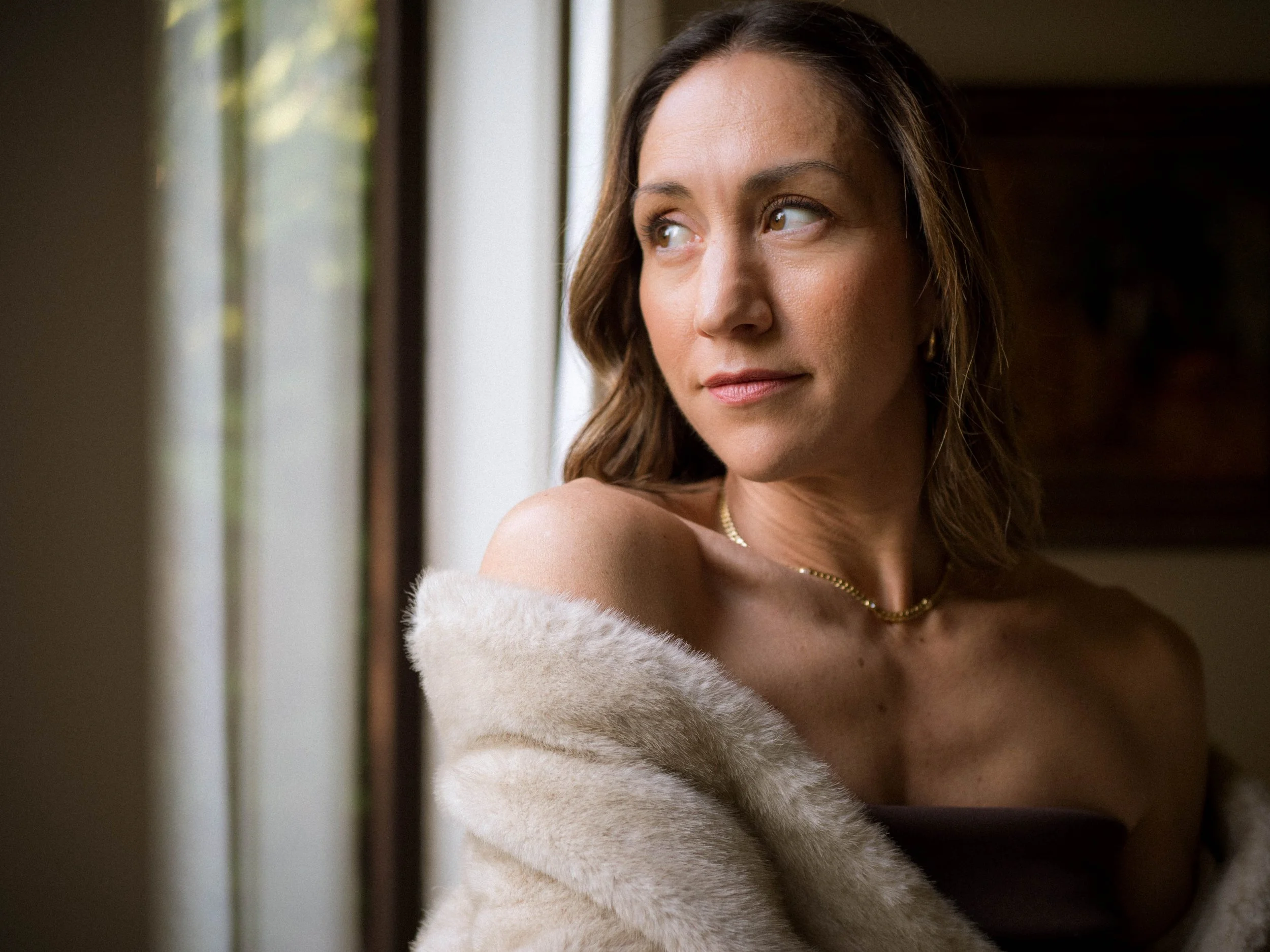 A woman looking out a window, wrapped in a beige fur coat, with a soft smile and wearing a gold necklace.