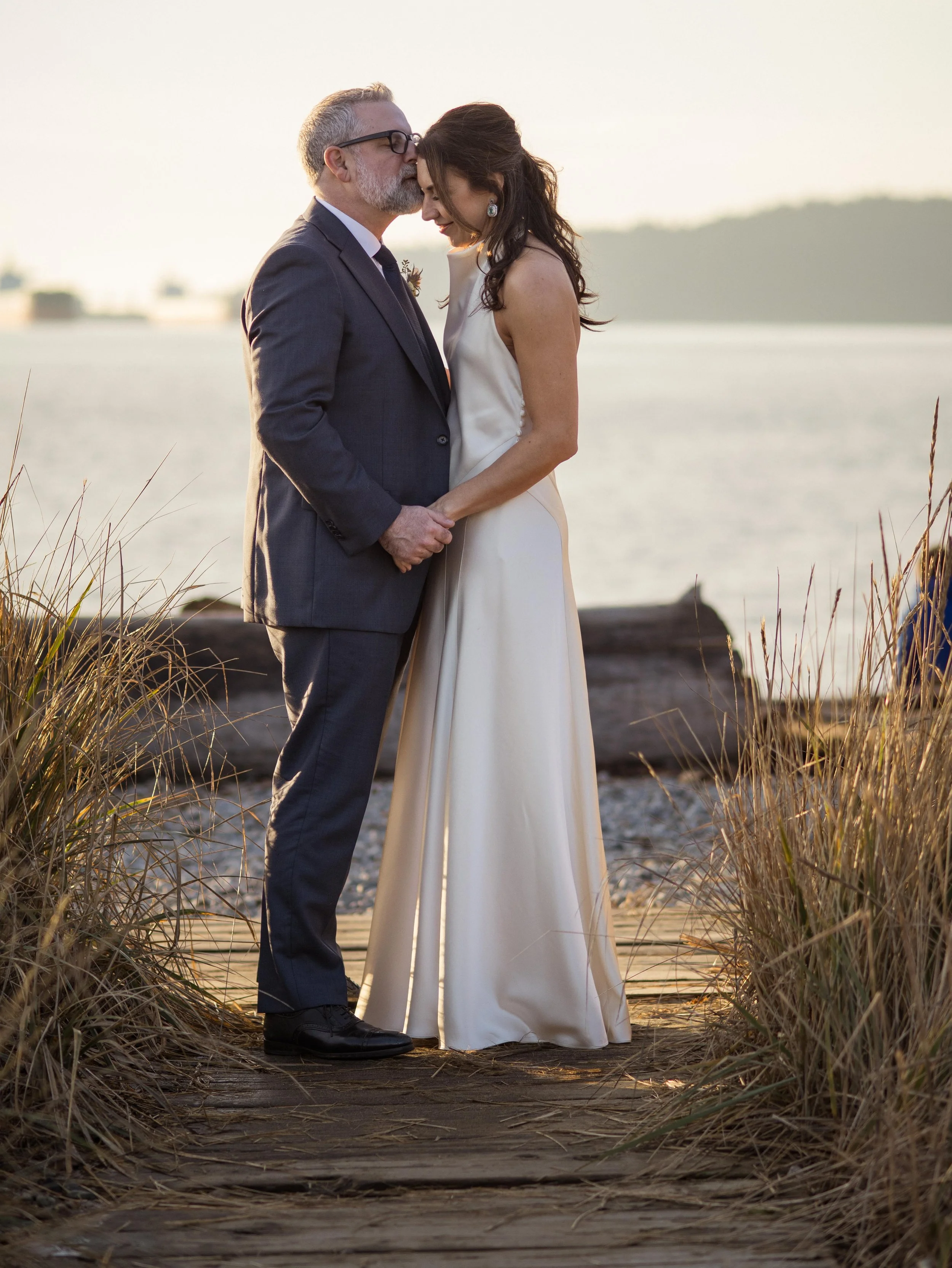An older man in a gray suit, kissing a woman in a white satin dress on her forehead by a body of water at sunset.