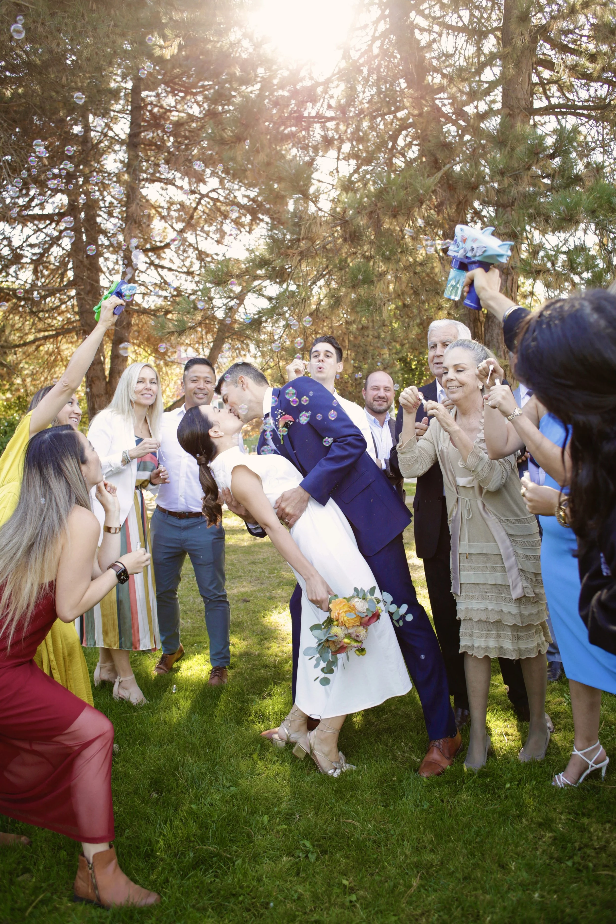 A couple kissing at their wedding surrounded by friends and family outdoors with sunlight filtering through trees, some people blowing bubbles and celebrating.