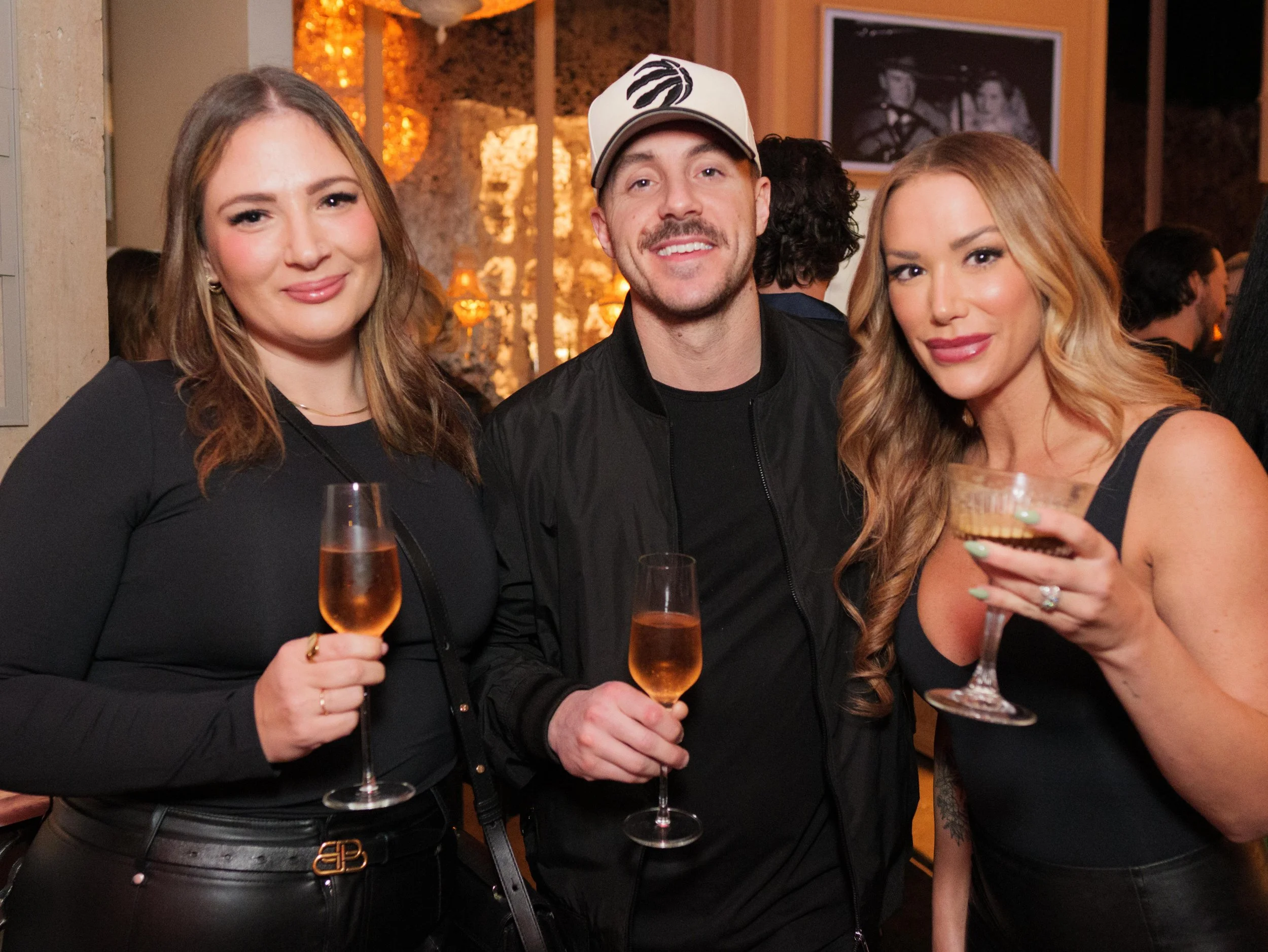 Three people, two women and one man, are at a social gathering holding glasses of champagne and cocktail, smiling at the camera.