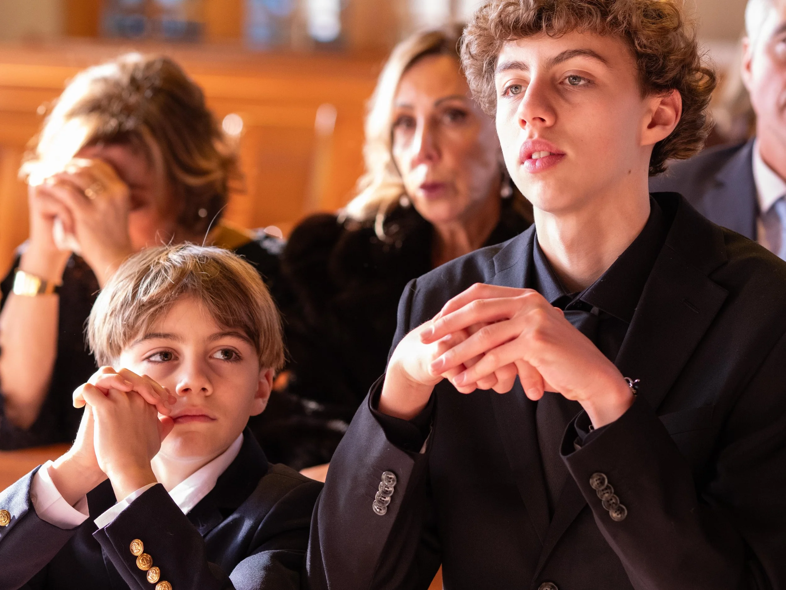 A group of people sitting in a formal setting, including two boys in the foreground wearing suits, with a woman in the background appearing concerned. The scene suggests a serious or emotional event.