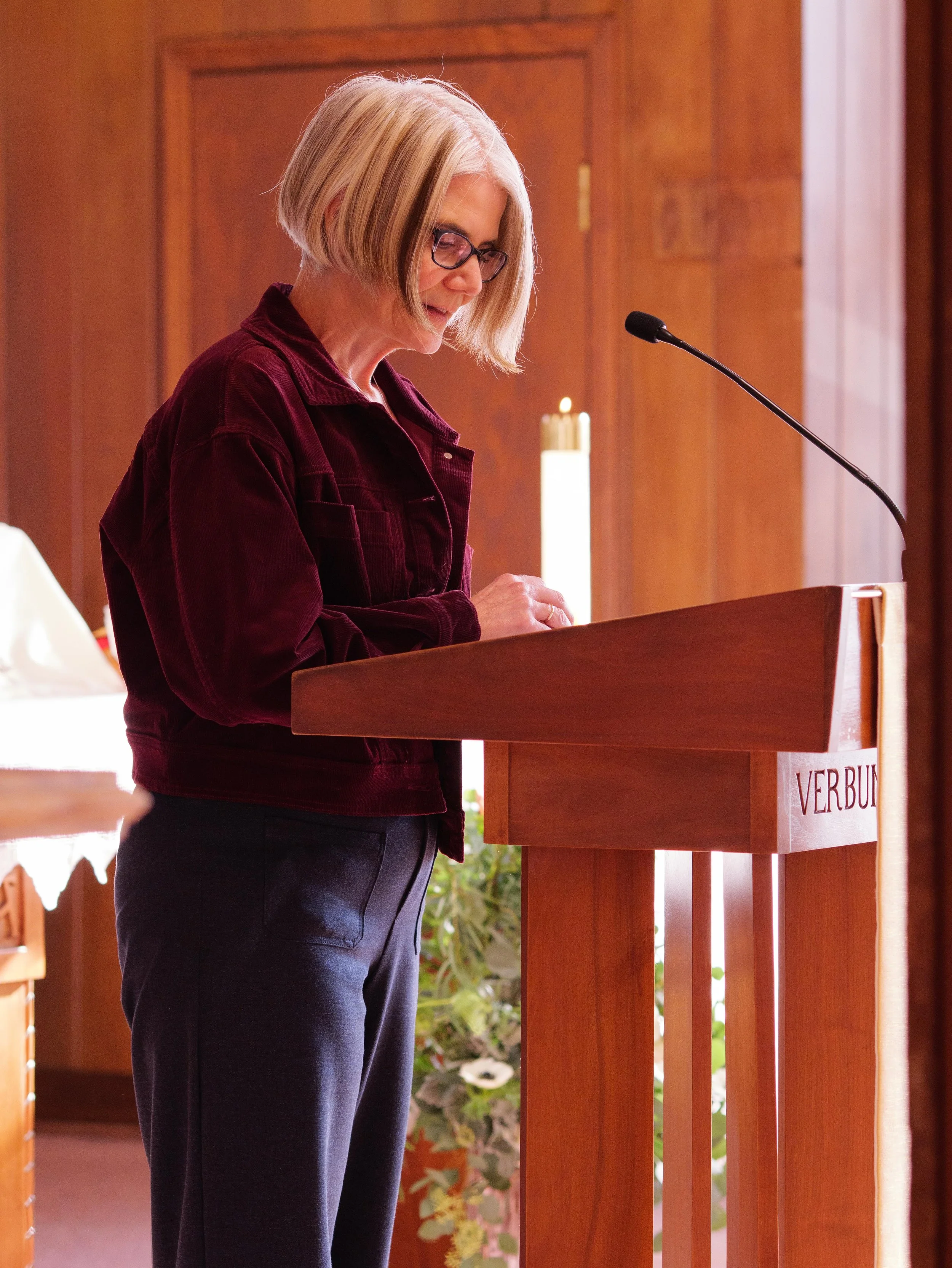 A woman with blond hair, wearing glasses, a burgundy jacket, and dark pants, is standing at a wooden lectern inside a wood-paneled room, reading or reciting from notes.