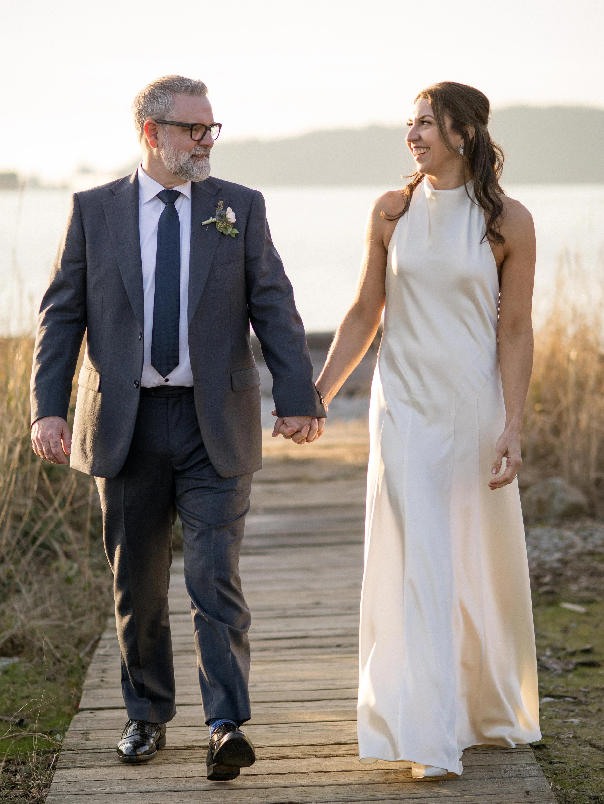 A man and woman, dressed in formal attire, walking hand-in-hand on a wooden pathway near a body of water during sunset.