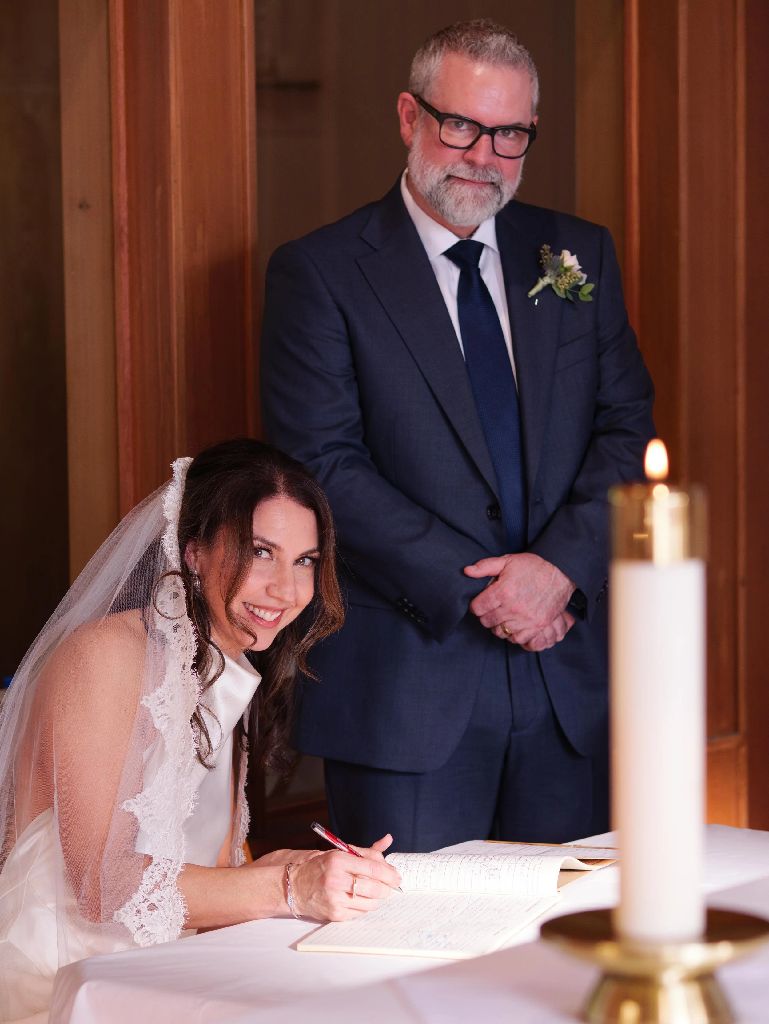A bride with long brown hair and a white lace dress signs a document at a wedding ceremony table, while a man in a dark suit and glasses stands nearby with his hands clasped, inside a wooden-paneled room.