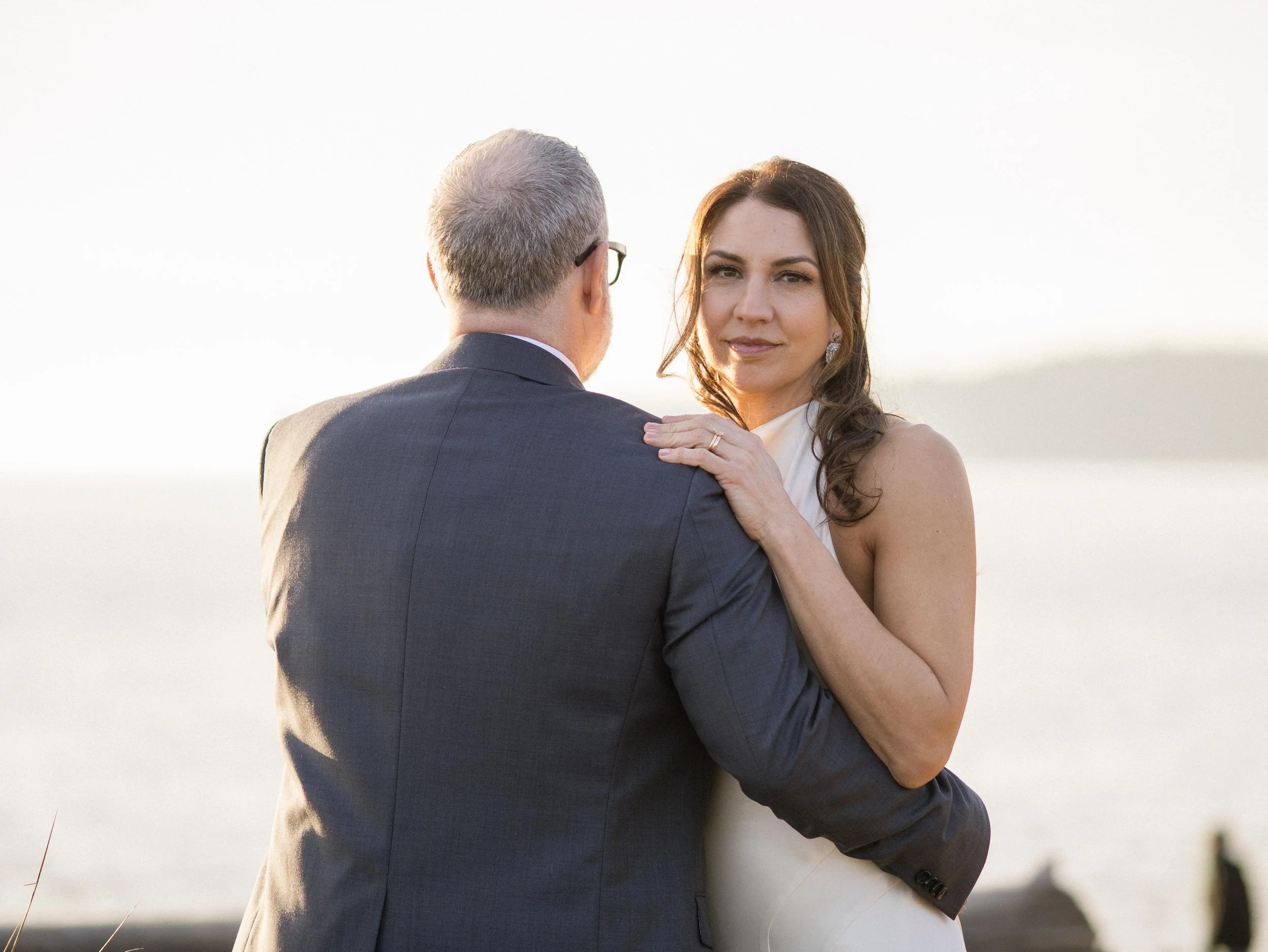 A couple dressed in formal attire, standing outdoors near a body of water at sunset, embracing each other.