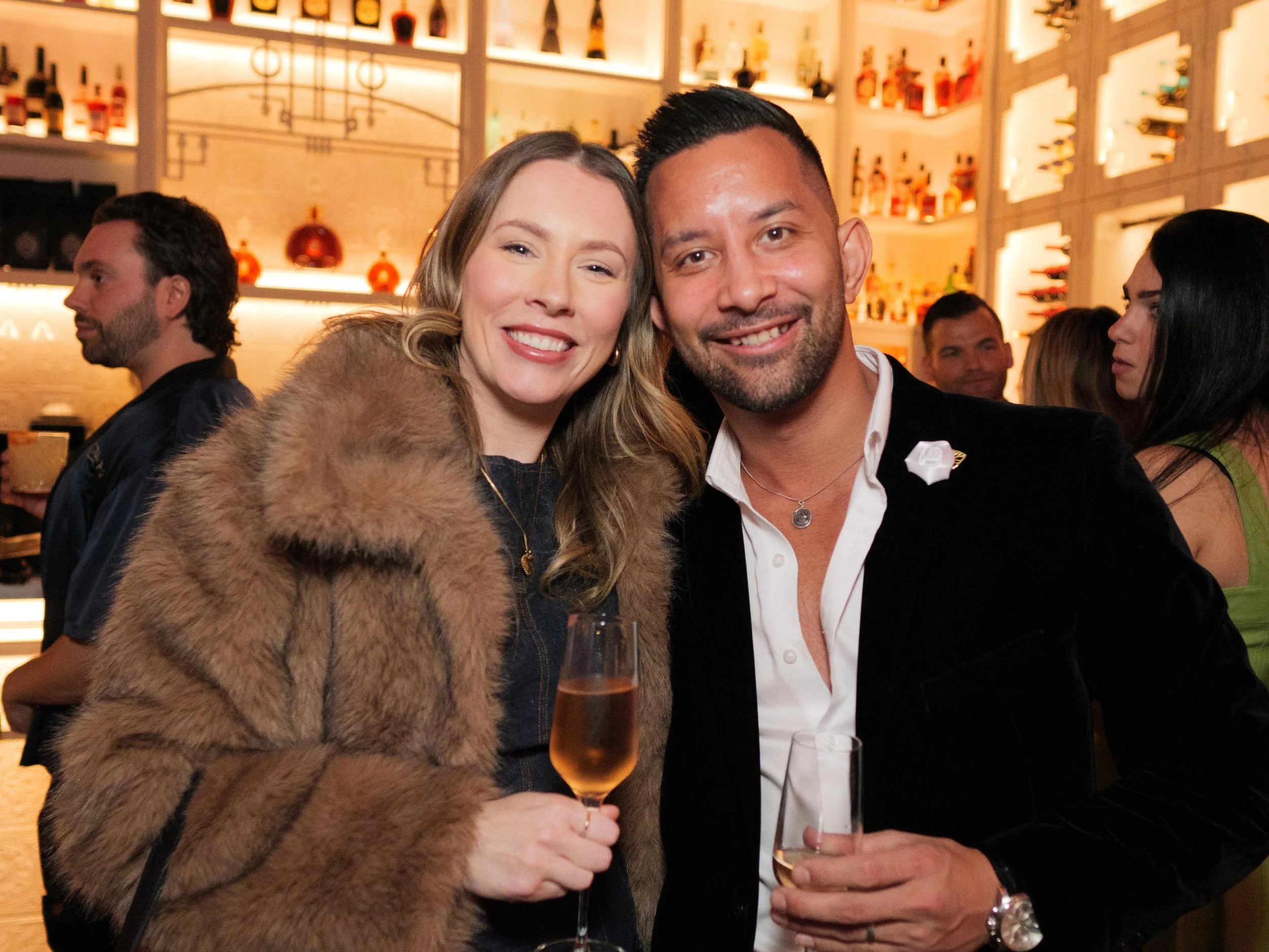 A smiling woman and man holding glasses of champagne at a social event or party, with a bar in the background.