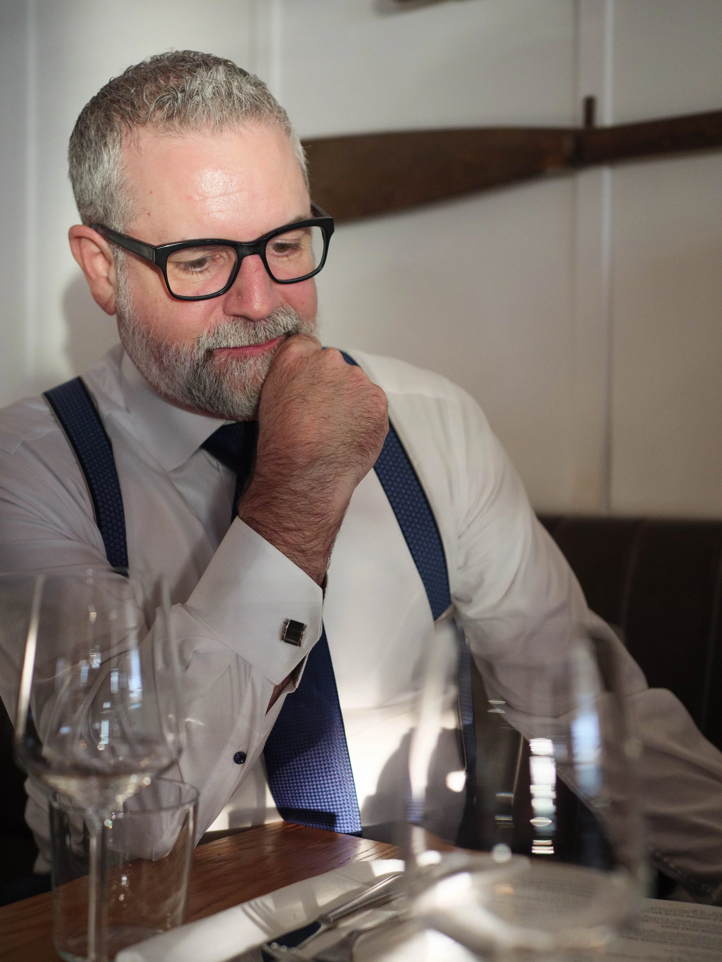 A man with gray hair and beard wearing black glasses, a white shirt, and suspenders sitting at a table in a restaurant, with a contemplative expression.