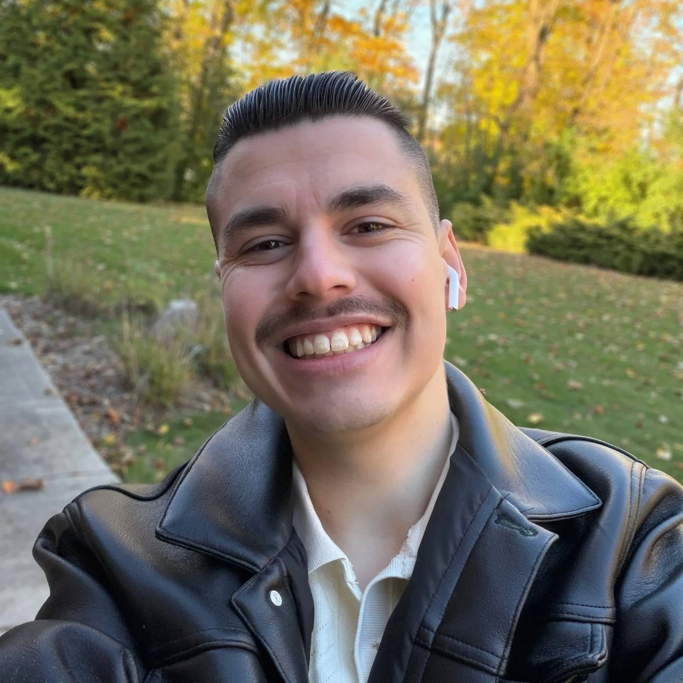 A smiling young man with short, dark hair styled upward, wearing a leather jacket, white shirt, and white wireless earbuds, taking a selfie in a park during autumn with trees and fallen leaves in the background.