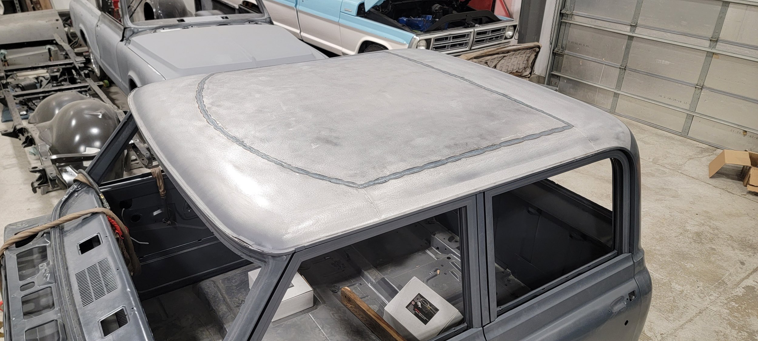 Vehicle in a workshop with a metallic roof patch and unfinished interior, surrounded by car parts and other vehicles.
