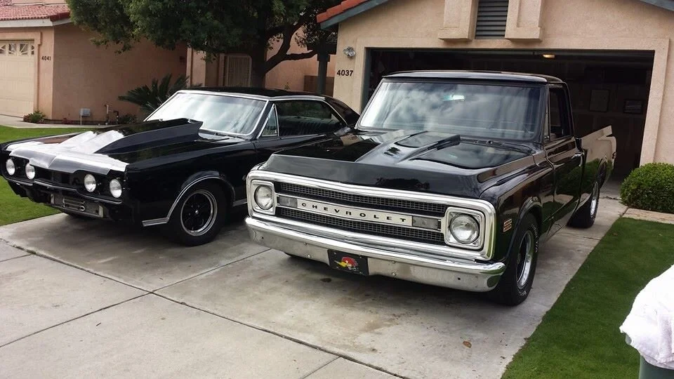 Two classic black vehicles parked in a driveway, a vintage Chevrolet truck on the right and a classic muscle car on the left, in front of a house with a garage.