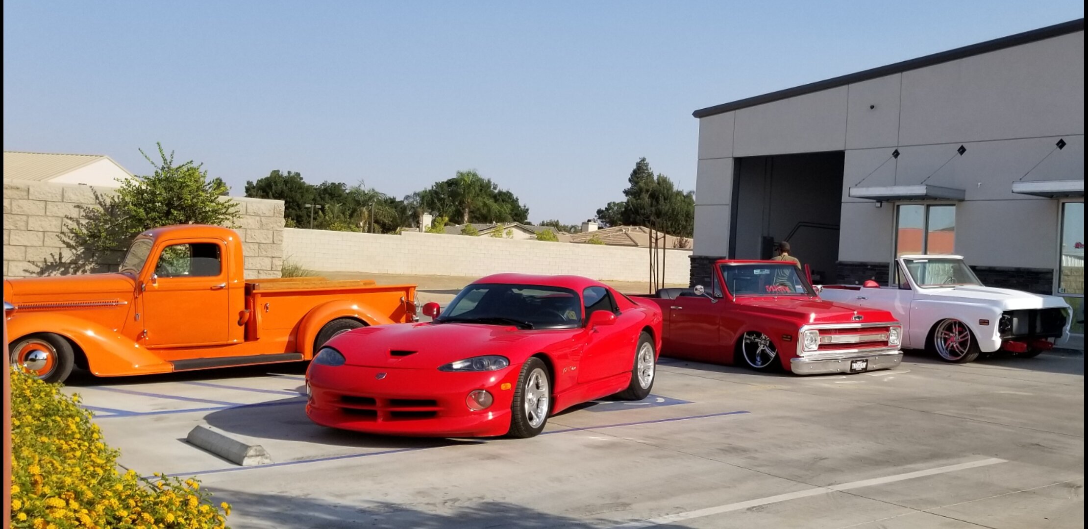 Classic and vintage cars parked outdoors, including an orange pickup truck and two red convertibles, with a building in the background.