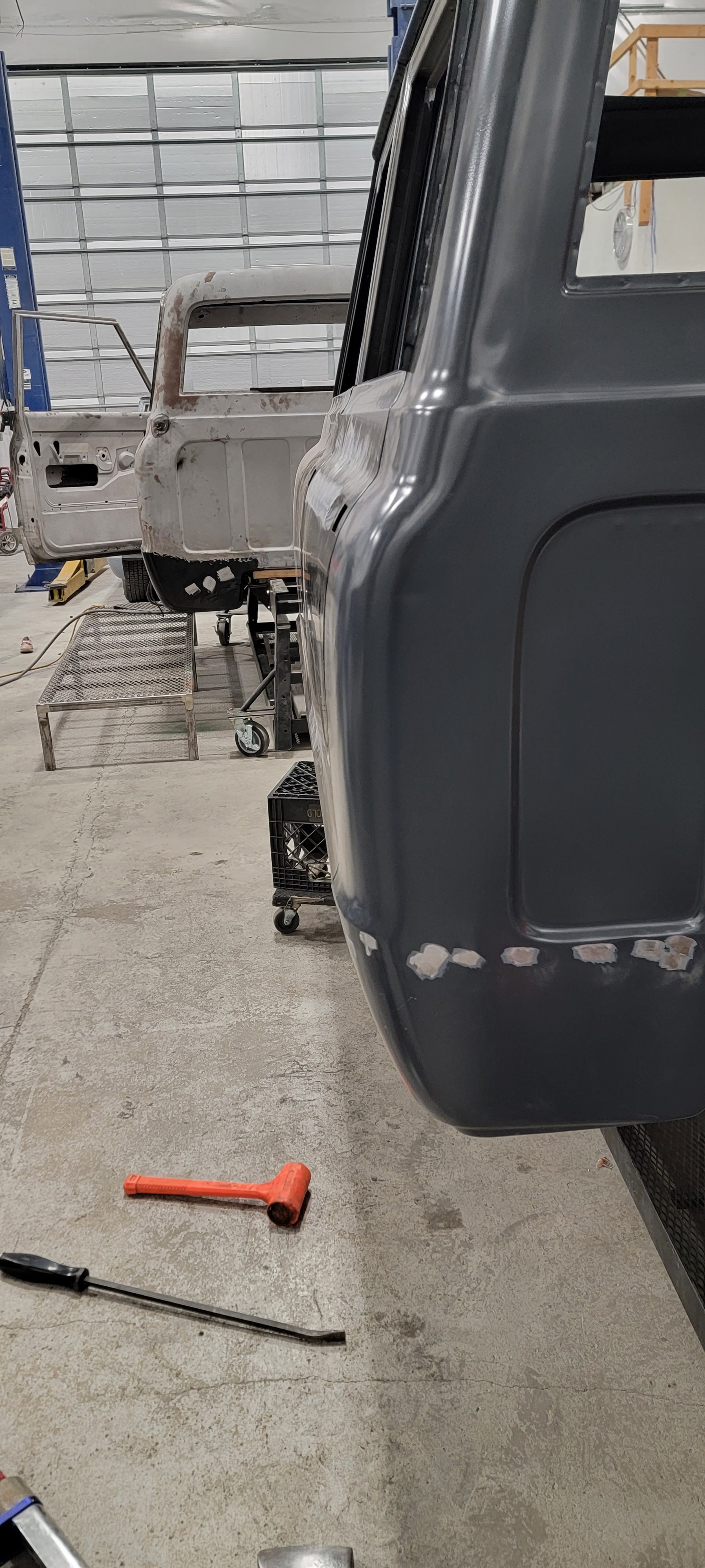 Workshop with two vehicle bodies, one gray and one primed, undergoing restoration, with tools on the floor.