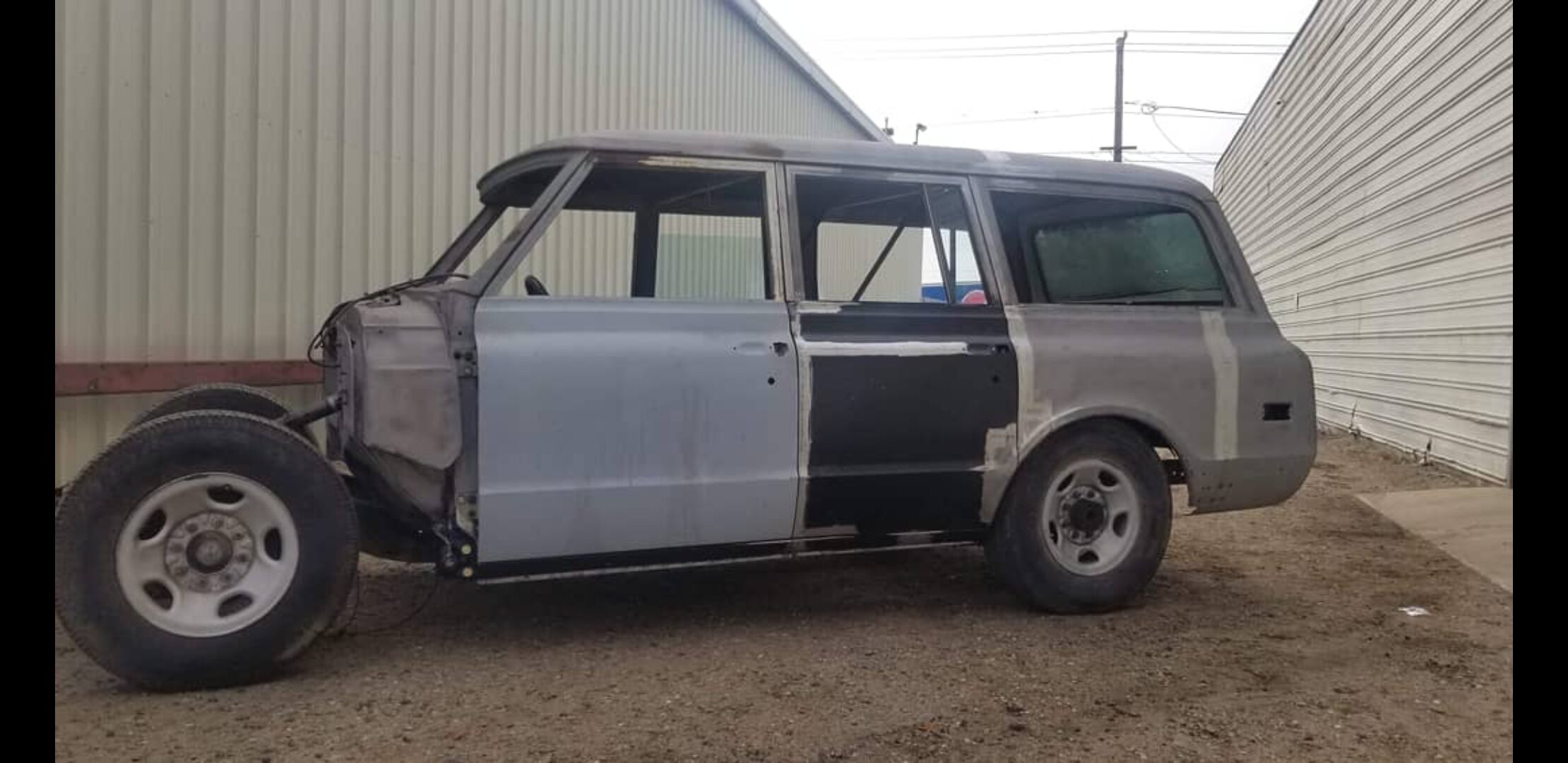 Partially restored vintage SUV without front end parked beside metal buildings.