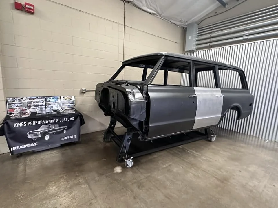 Car restoration workshop with a classic wagon frame on a rolling stand, adjacent to a table displaying automotive images and a Jones Performance & Customs banner.