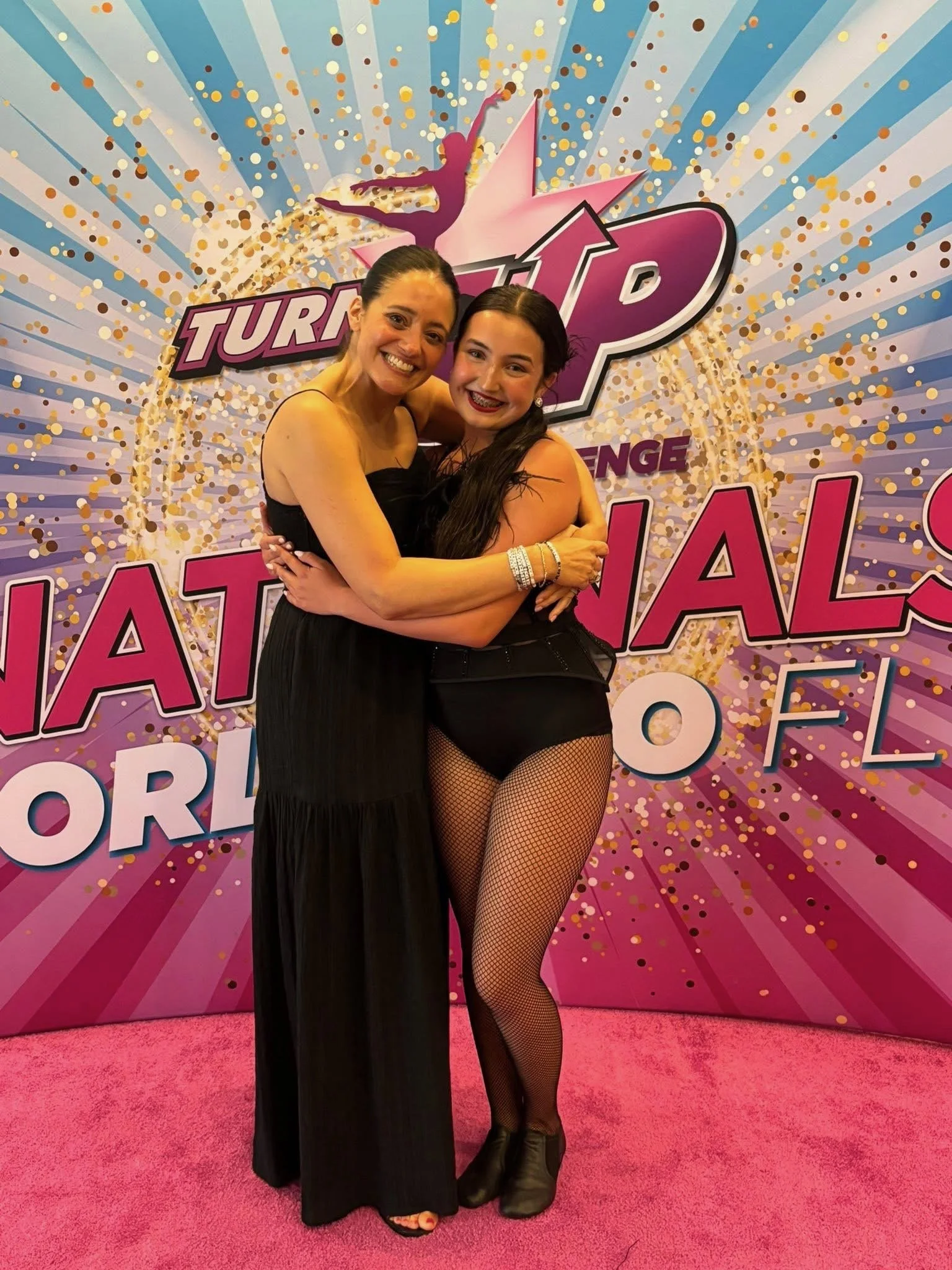Two women embracing and smiling at the camera in front of a colorful backdrop with the words "Turn Up" and "National" visible, at a dance or talent competition event.