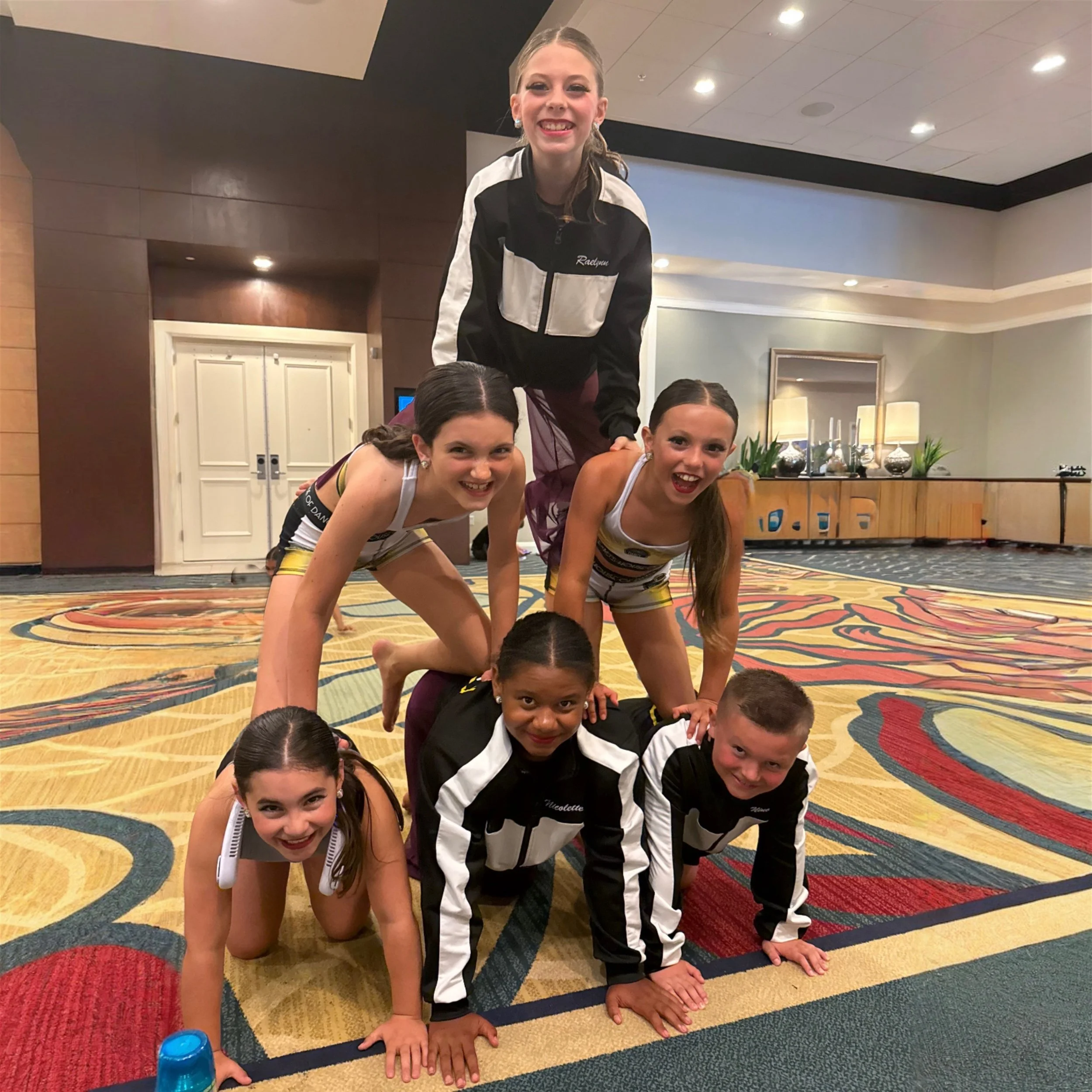 Group of six young cheerleaders forming a human pyramid in an indoor setting with colorful carpet and decorative background.