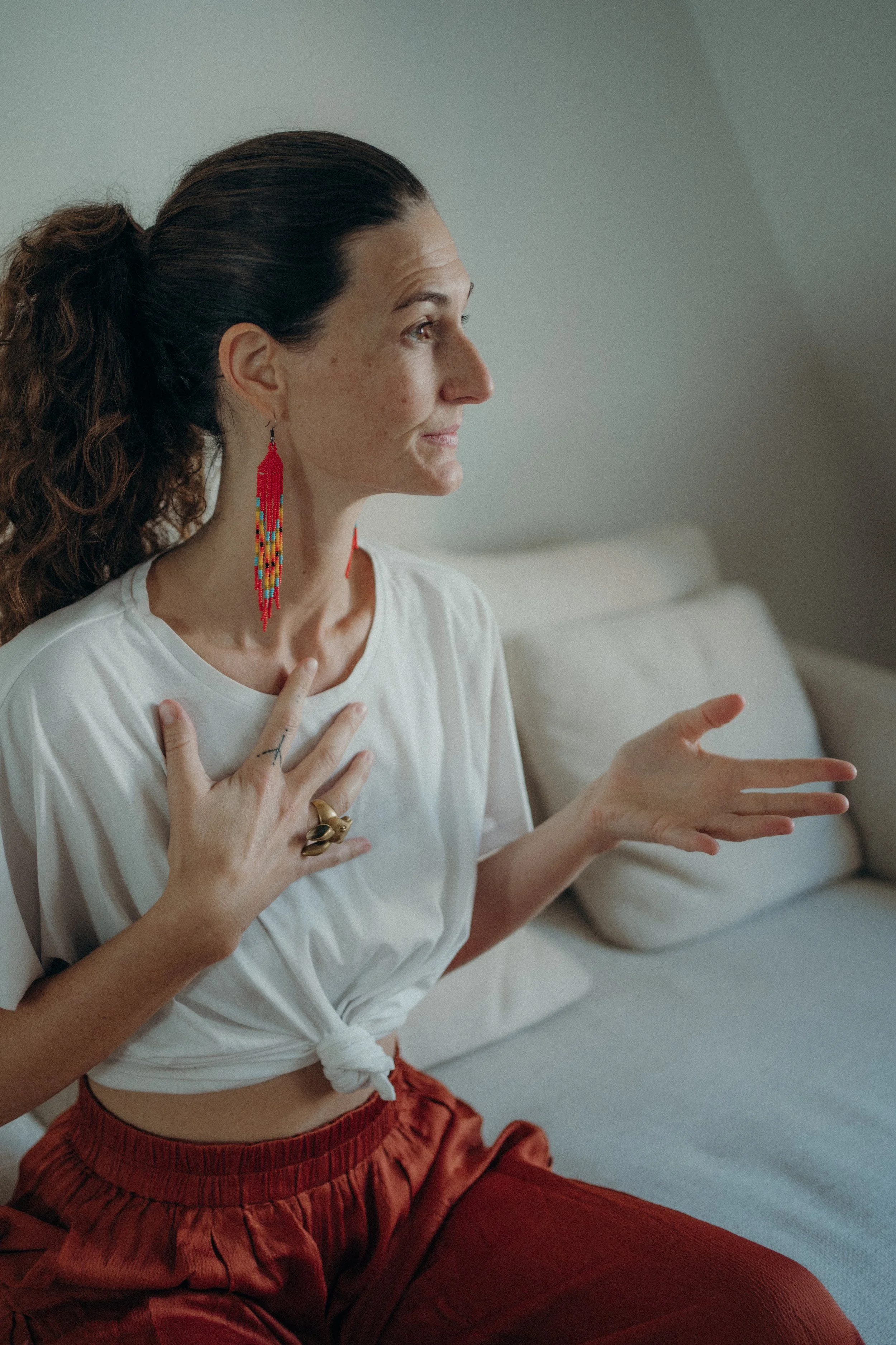 Woman with long hair wearing a white shirt and red pants, sitting on a couch, gesturing with her hands. She has colorful beaded earrings and a large ring on one hand.