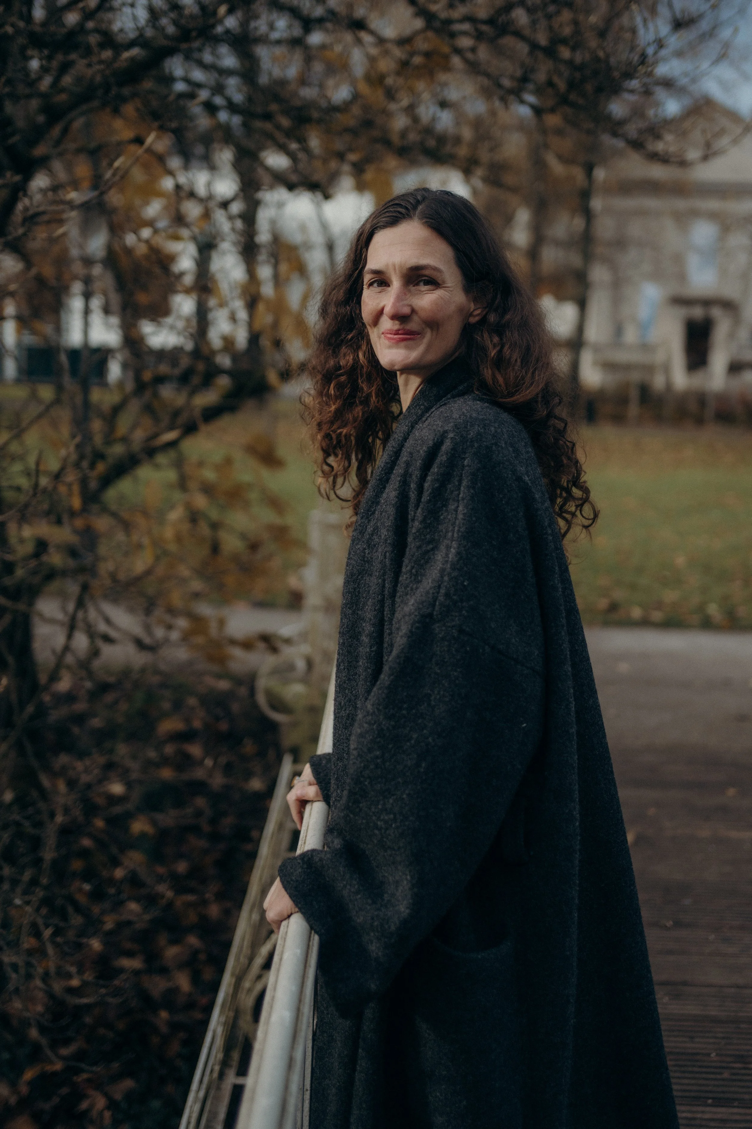 Woman with long curly hair smiling outdoors in a gray coat, standing on a wooden bridge with autumn trees in the background.