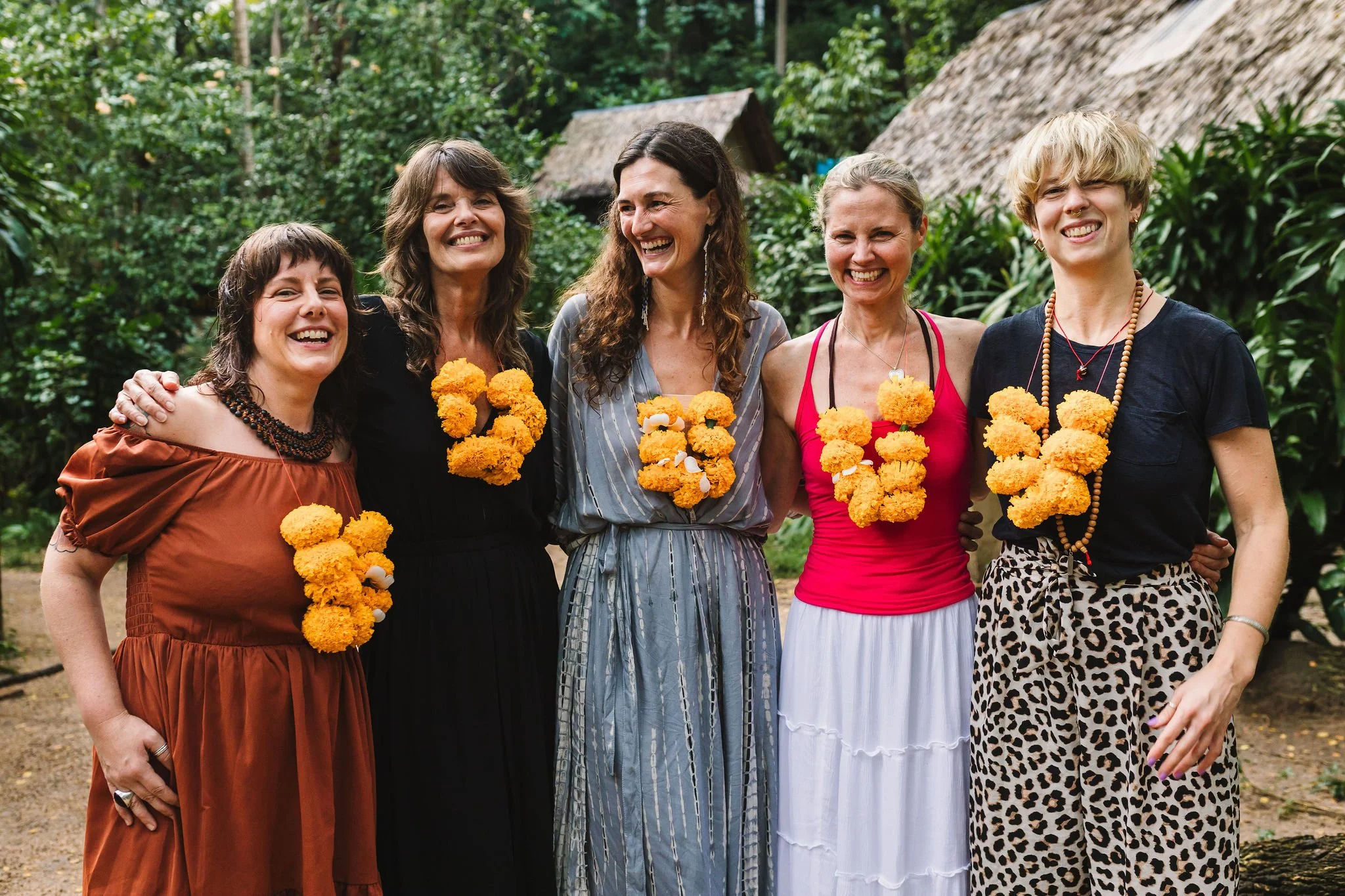 Five women standing together outdoors, each wearing a garland of orange flowers. They are smiling and appear to be in a natural setting with greenery and thatched-roof huts in the background.