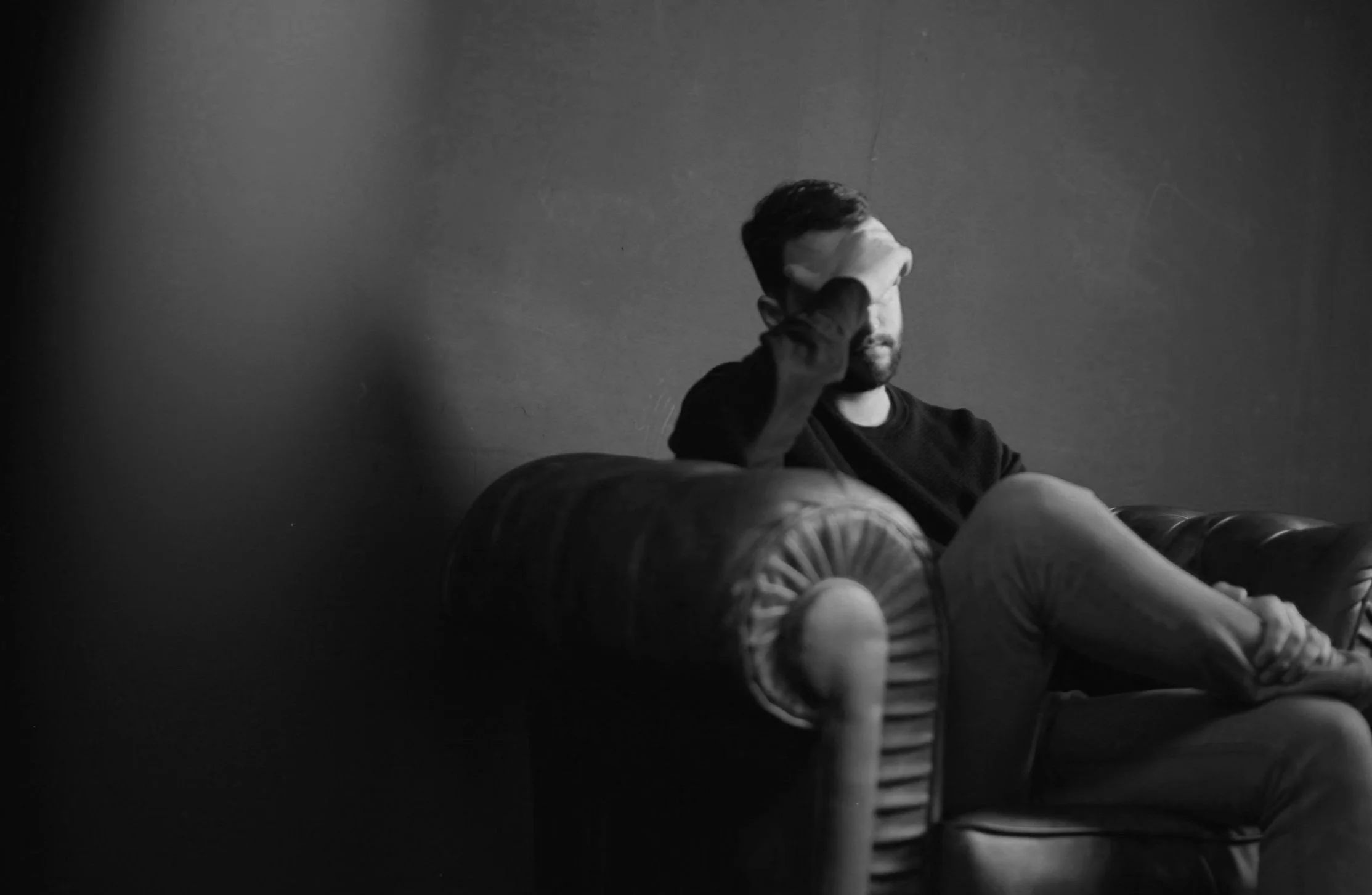 A man sitting on a vintage sofa with a distressed posture, head resting on his hand, in a black and white photo.