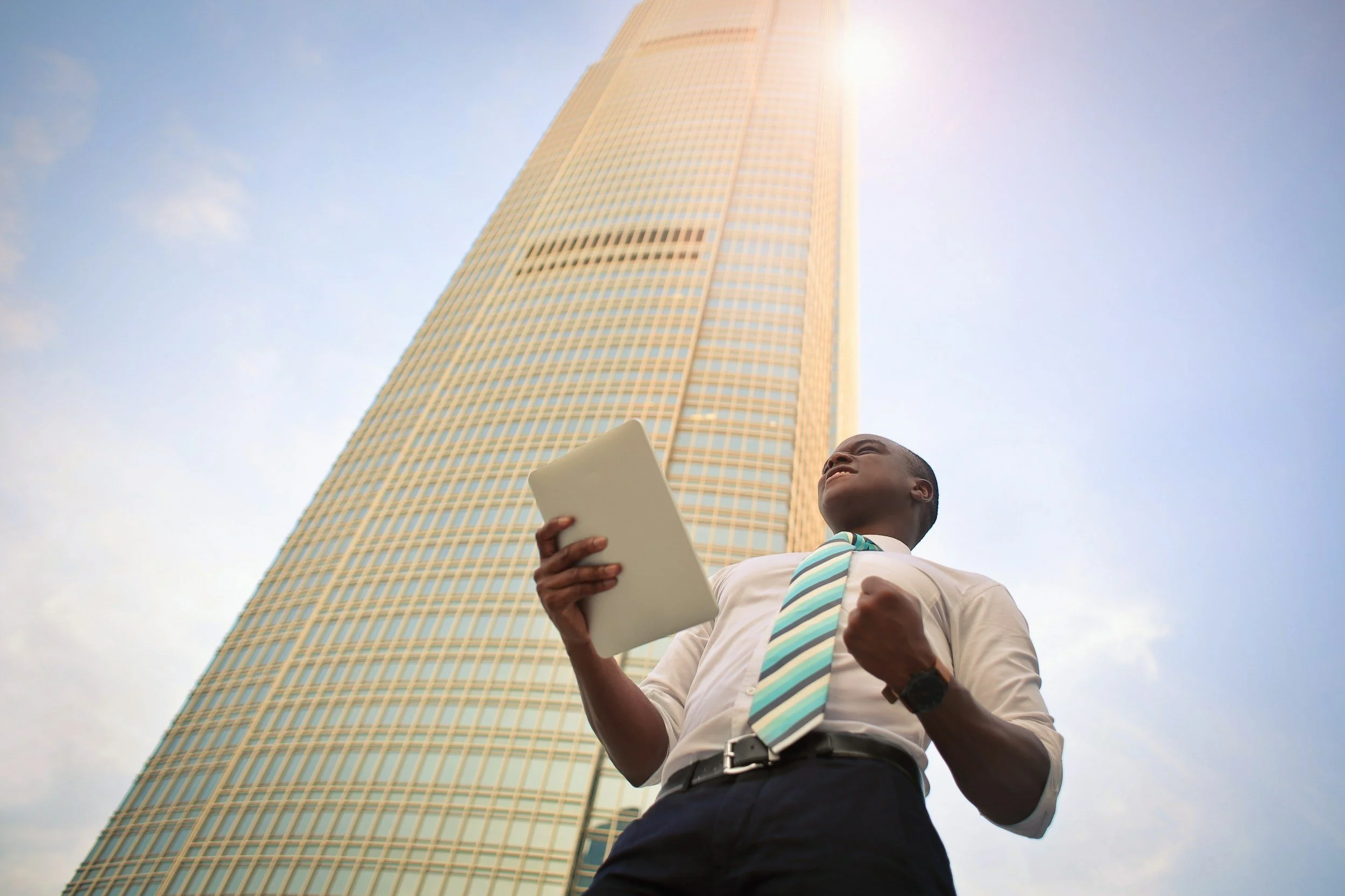 Confident businessman in a white shirt, striped tie, and black pants holding a tablet and making a fist while standing in front of a tall skyscraper on a sunny day.