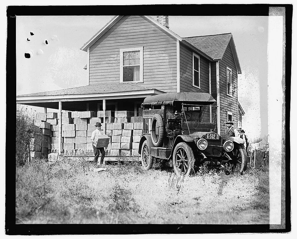 Black and white photo of a vintage car parked in front of a two-story house with a front porch, and three men wearing hats standing nearby; one man is holding a box, and another is near the car.