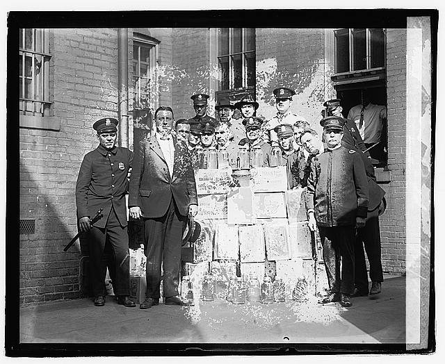 Group of police officers and children standing behind a table with boxes, outside brick building, black and white photo