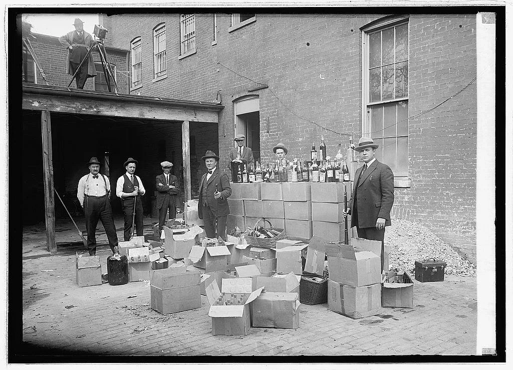 A black and white photograph of men standing outside a brick building, with boxes and bottles of alcohol on display, some open and some in cases, suggesting a sale or gathering during the early 20th century.
