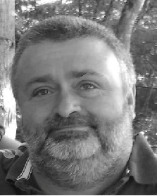 Black and white photo of a man with a beard and short hair, outdoors with trees in the background.