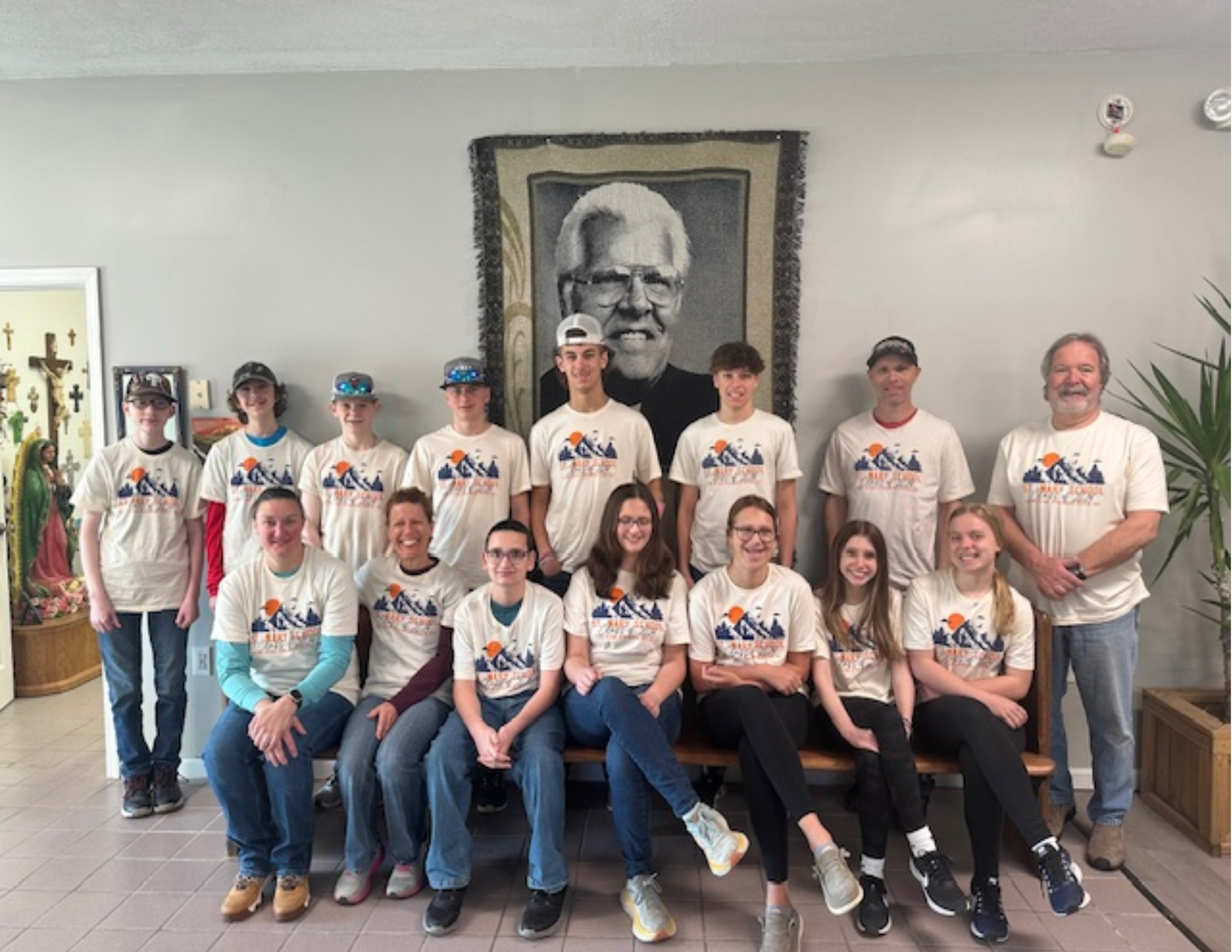 A group of young people and an adult man, all wearing matching white T-shirts with a mountain and sunset design, posing in front of a large portrait of an older man with glasses and white hair. The group is indoors, with religious items on the left and a plant on the right.