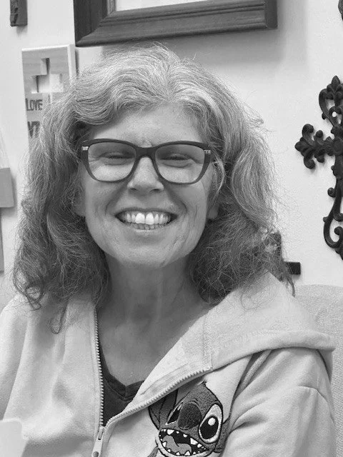 Smiling woman with glasses and curly hair, wearing a hoodie with a cartoon character on it, indoors with wall decorations.