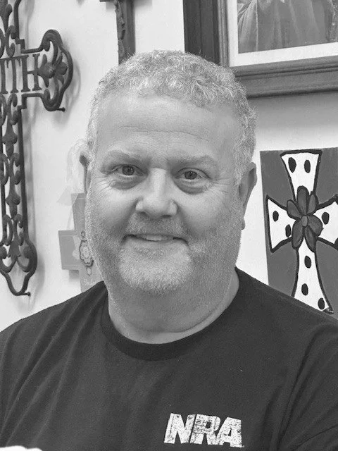 A man with curly gray hair and a beard smiling at the camera, wearing a black T-shirt with an NRA logo, standing indoors with various wall decorations and artwork in the background.