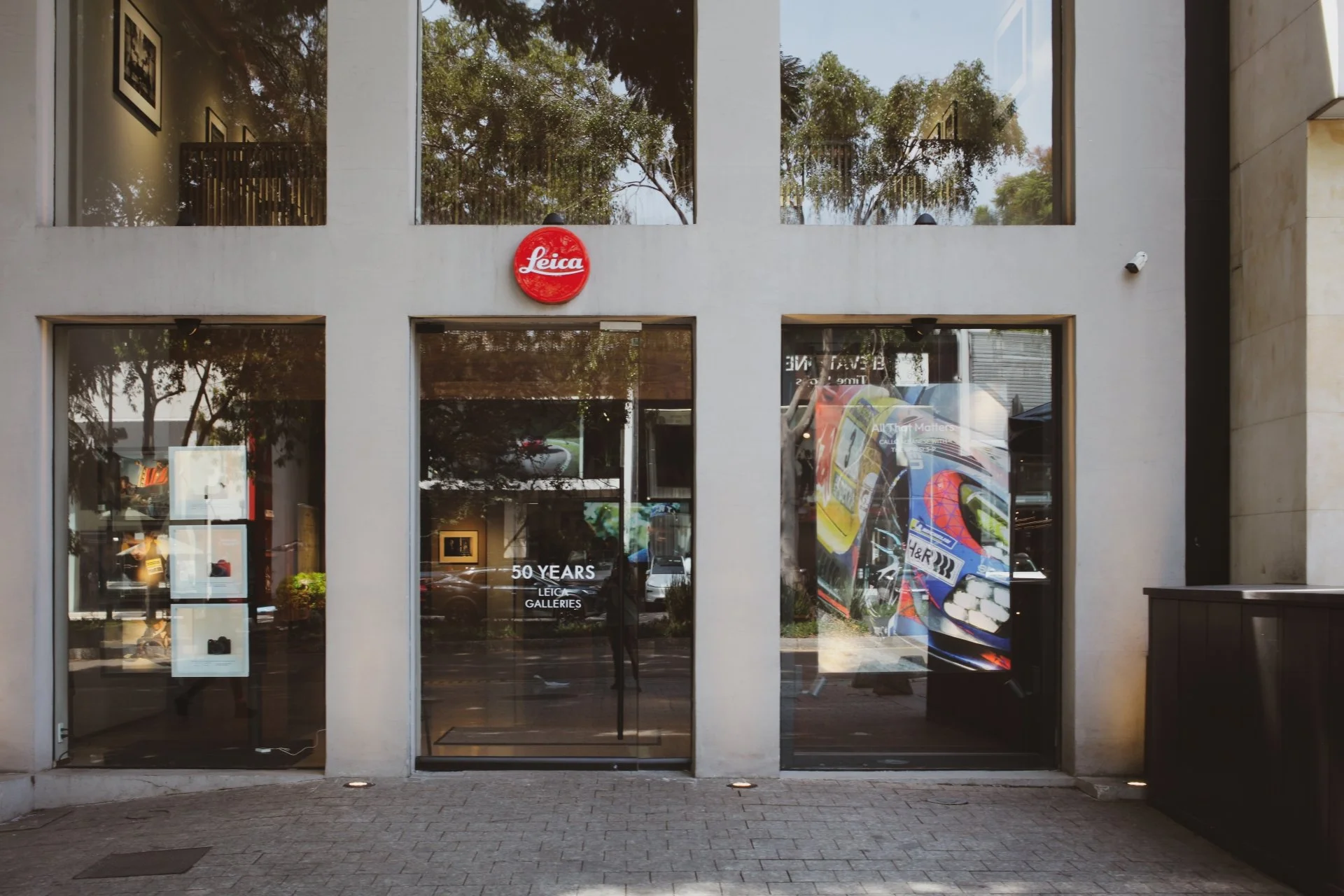 Front entrance of a building with large glass windows and doors, decorated with a red circular Leica sign above the door, reflecting the street scene and trees in the glass.