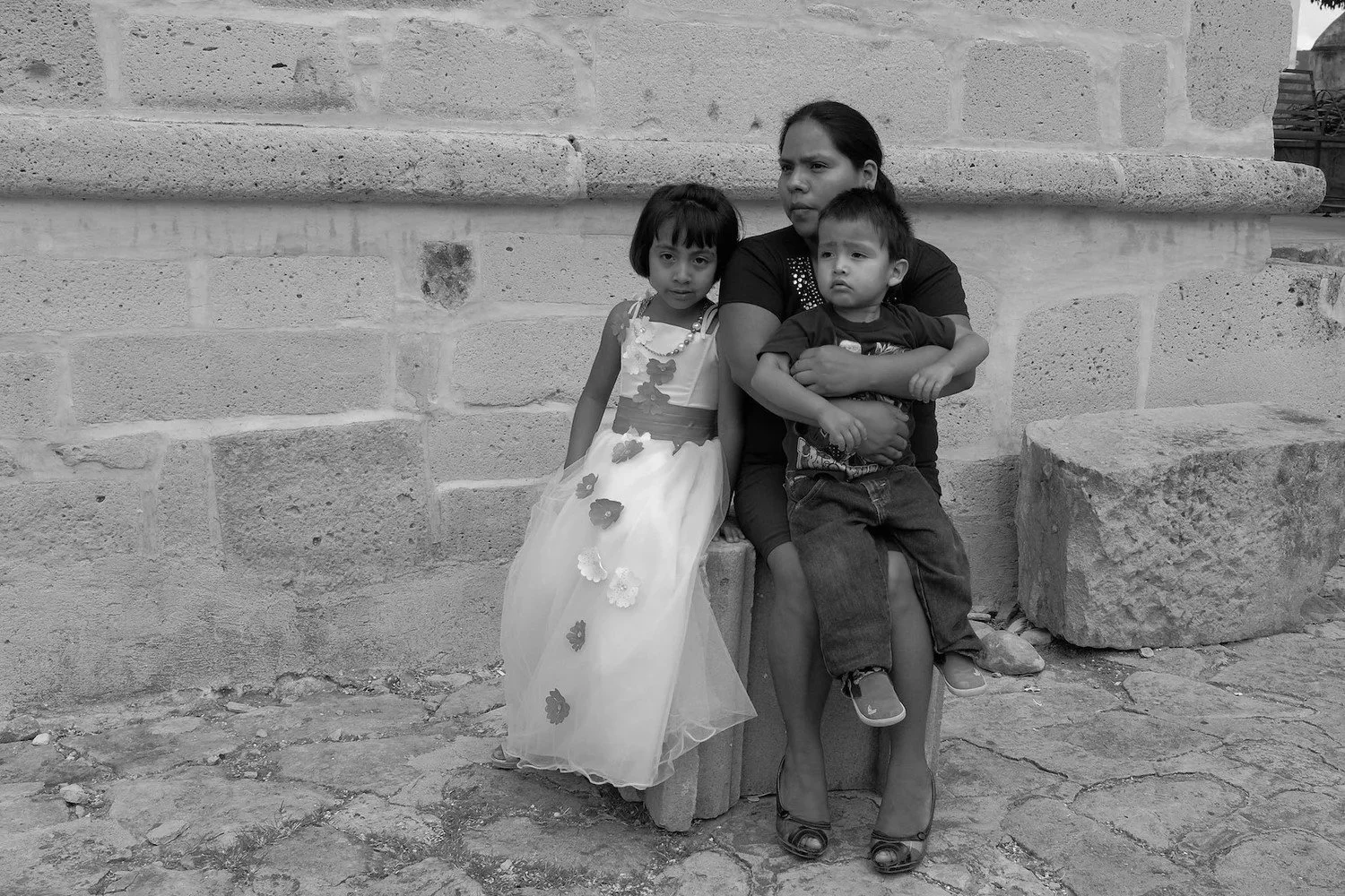A woman sitting on a bench with two children, one girl and one boy, against a stone wall. The girl is wearing a dress with floral decorations, and the boy is being held by the woman, wearing casual clothes. The photo is in black and white.