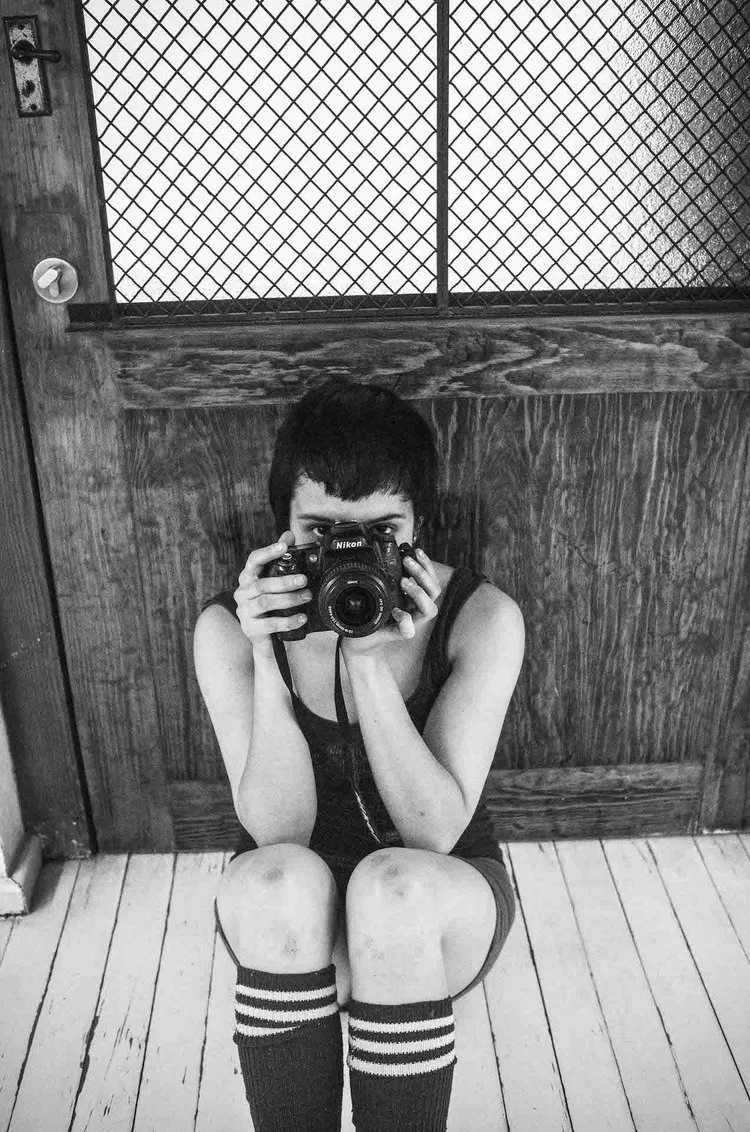 A person with short dark hair and sleeveless shirt sitting on a wooden floor, holding a camera in front of their face, taking a mirror selfie. The background features a wooden wall and a textured glass window.