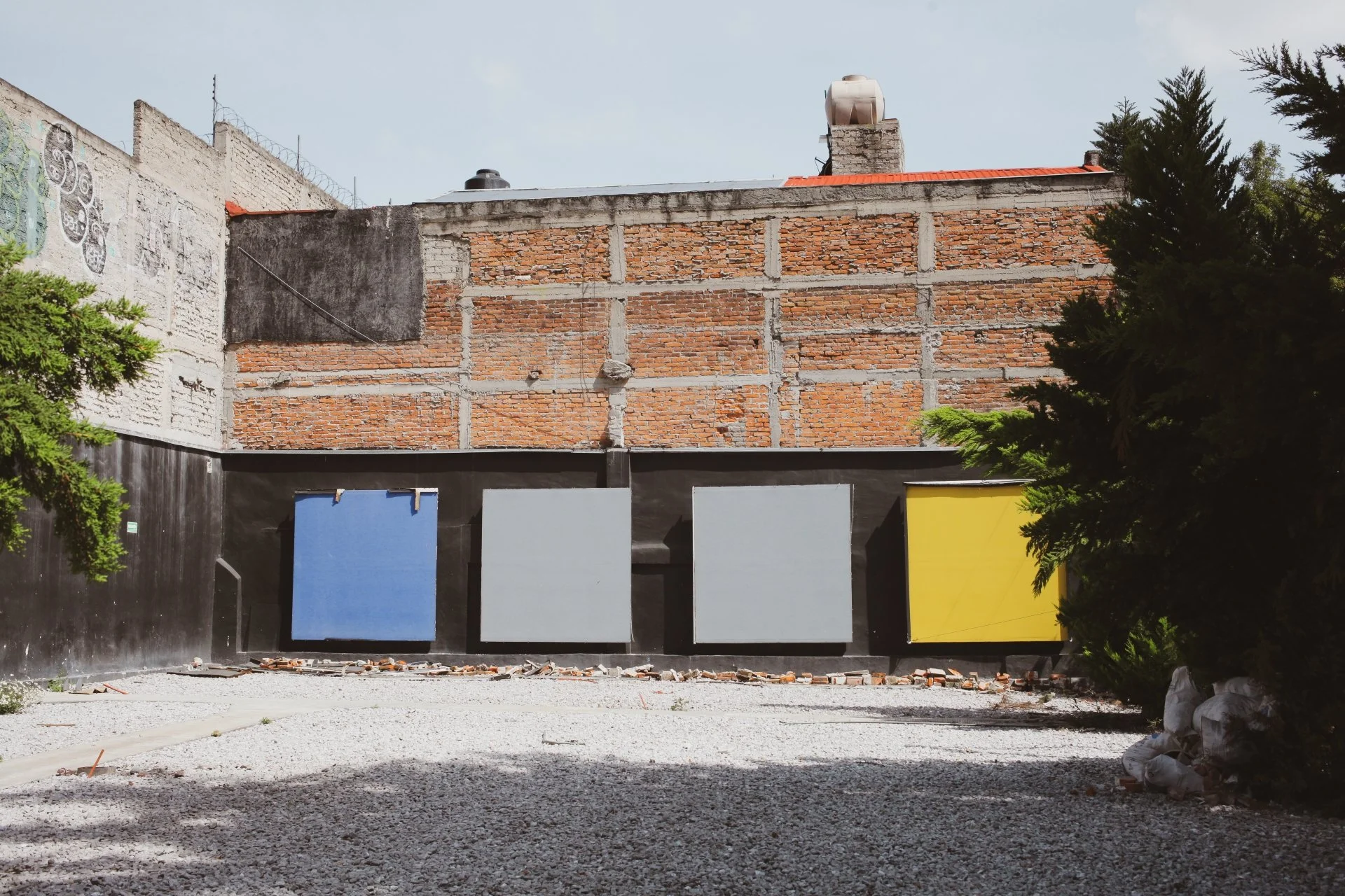 Building under construction with brick and concrete wall, colorful panels on the bottom, trees on the sides, and construction materials on the ground.