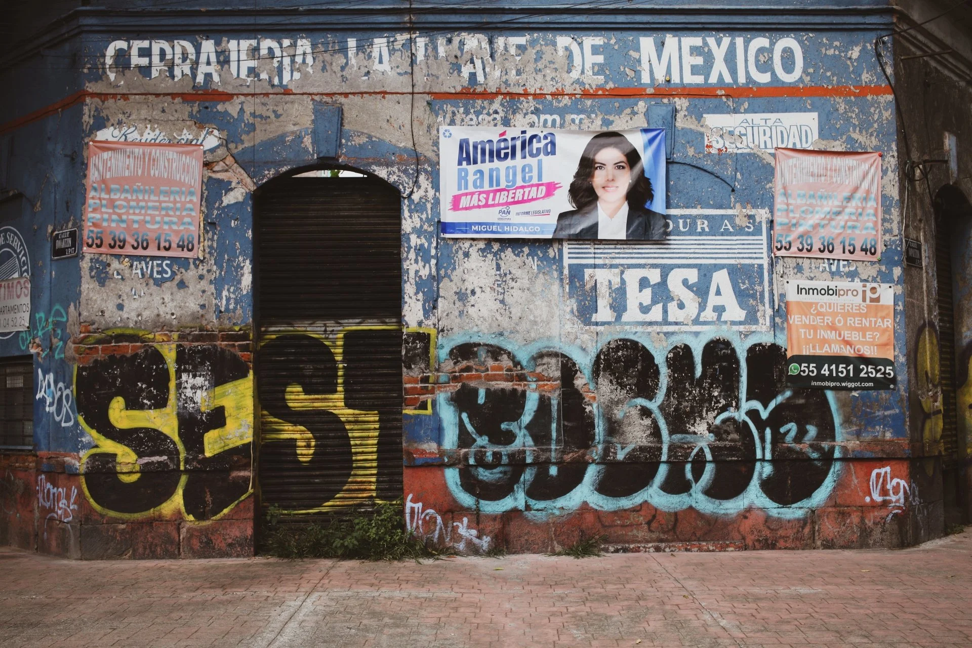 A wall covered with peeling paint and graffiti, with a large arch-shaped opening, adorned with political posters and advertisements, including a prominent campaign poster of a woman with dark hair, and various smaller signs for construction and real 