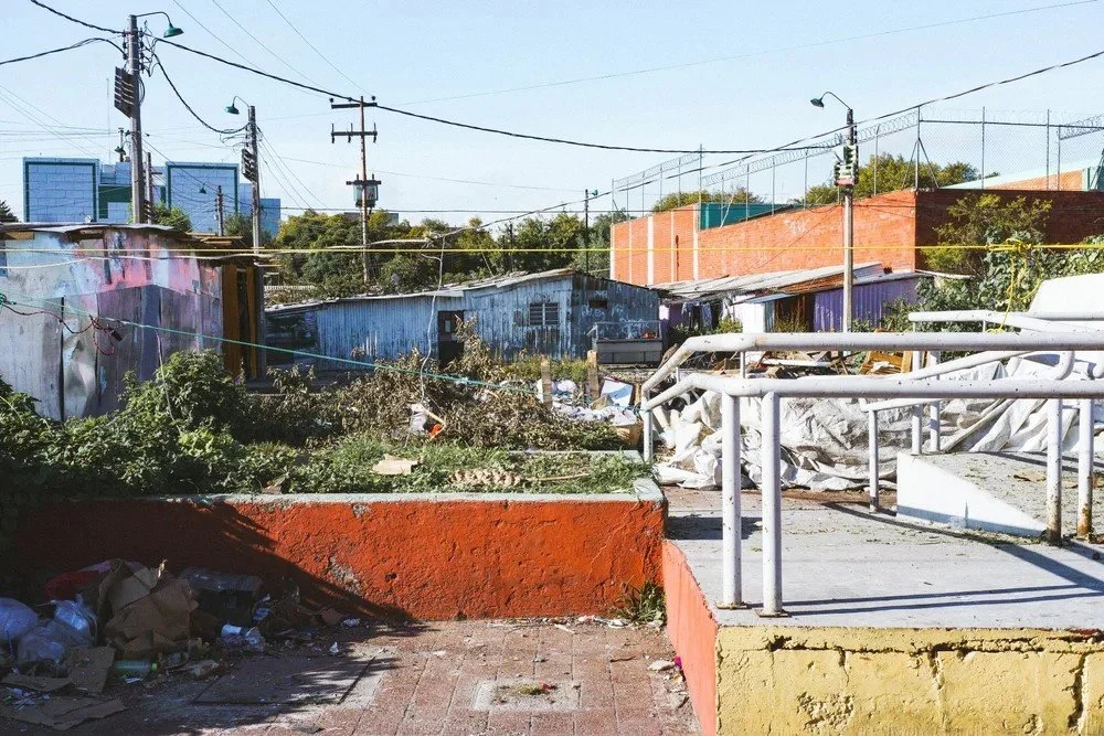 A neglected urban area with overgrown plants, scattered trash, and makeshift buildings made of metal and wood, with utility poles and wires overhead.