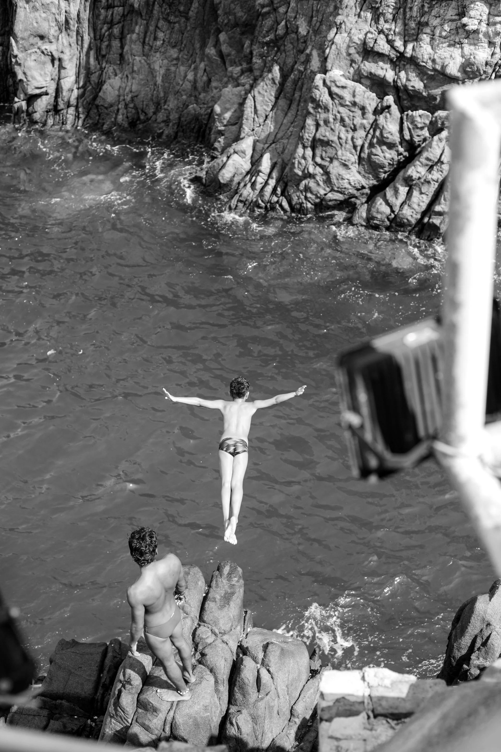 A person is mid-air, diving into the water from rocks along a rocky shoreline.