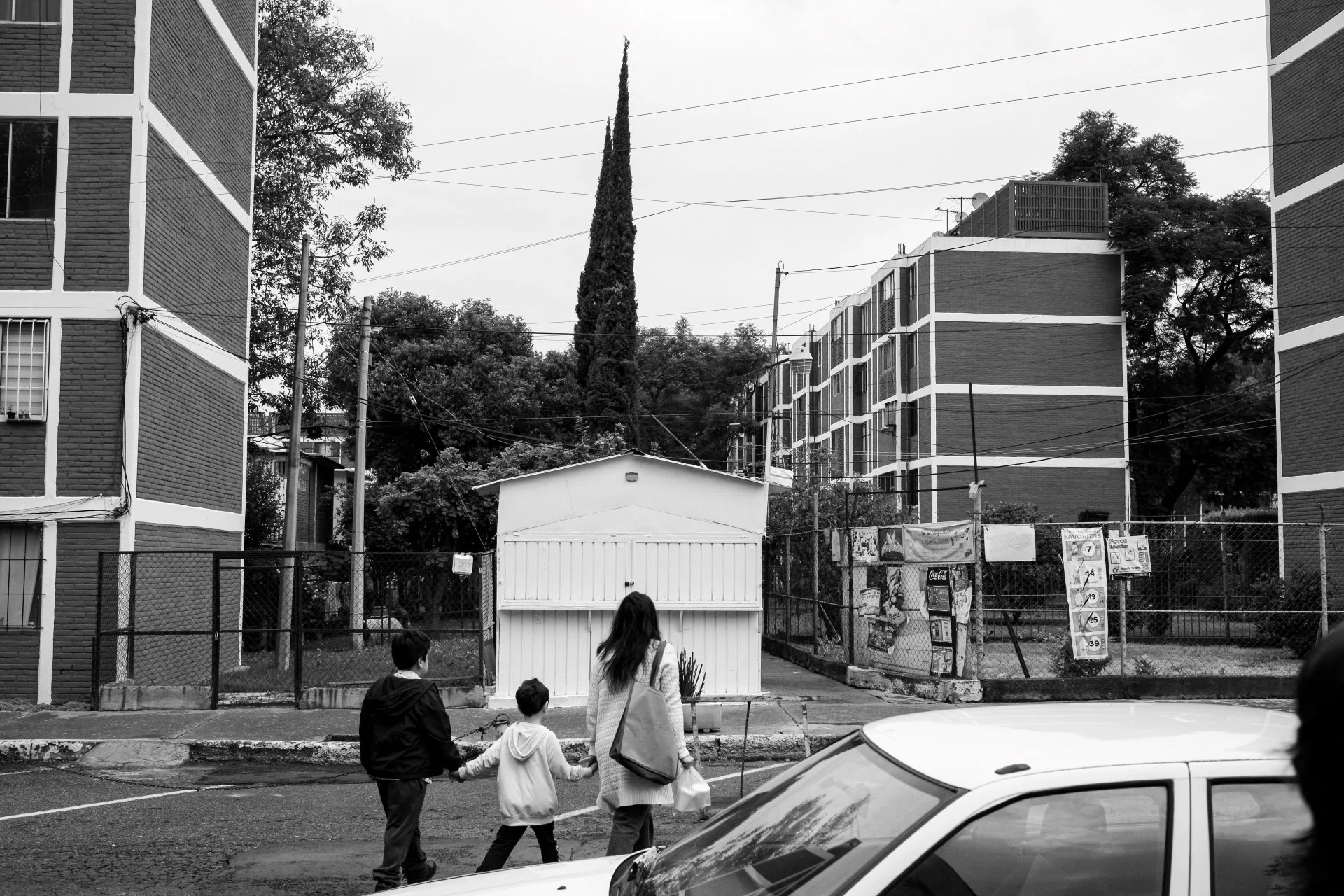 Kids and an adult walking across a street in front of a small white building. Behind them are tall apartment buildings, trees, and power lines. The scene is in black and white.