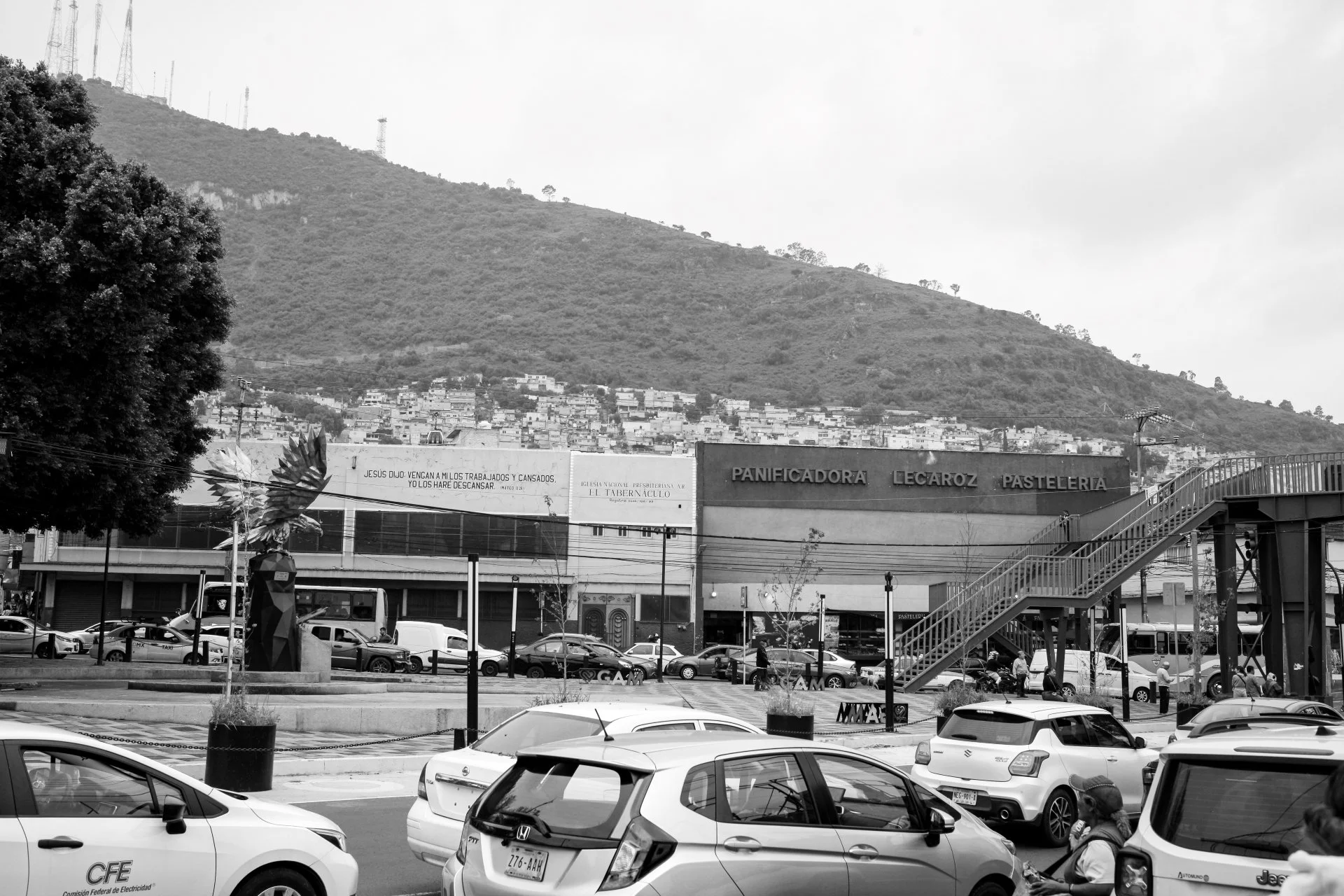 Black and white photo of a busy parking lot with various cars, a pedestrian staircase to a building labeled 'Panificadora Lecaroza Pasteleria,' a tree on the left, and a hillside in the background with numerous houses and radio towers.